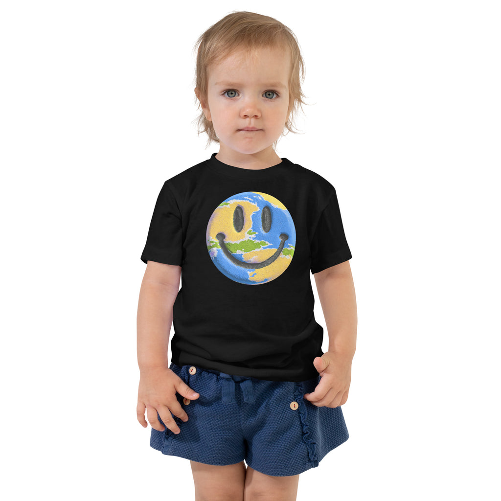 Product mockup. Toddler standing, wearing a black t-shirt with a blue, yellow, and green smiley face centered on the chest.
