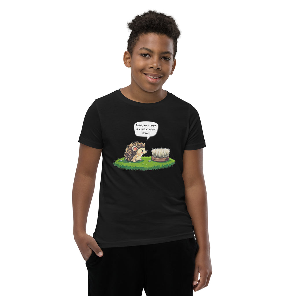 Product mockup. A boy wearing a black t-shirt featuring a playful cartoon illustration on the chest. The design shows a cute hedgehog standing on grass and looking at a scrubbing brush, with a speech bubble above the hedgehog that says, 'DUDE, YOU LOOK A LITTLE STIFF TODAY!