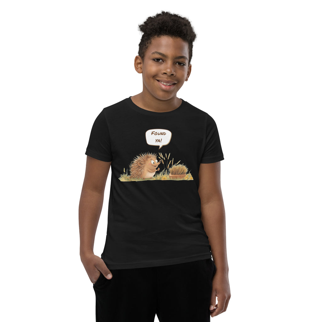 Product mockup. A smiling child wearing a black t-shirt printed with a playful illustration of a cheerful hedgehog popping up from the grass and saying “Found ya!” in a speech bubble, beside a second hedgehog curled into a ball, all set against a clean studio background.