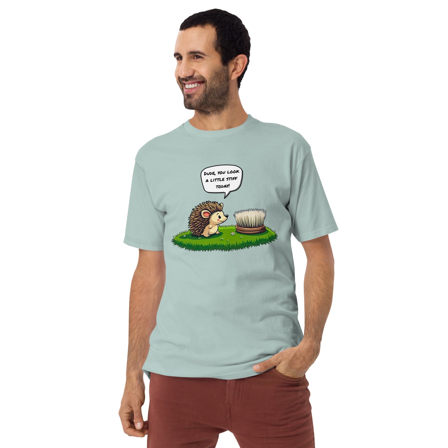 A bearded man with short dark hair wears a grabNade Men's Relaxed Heavyweight Tee in pale blue, featuring a cartoon hedgehog and hairbrush with the quote “Dude you look a little stiff today!” One hand is in his pocket. Premium cotton.