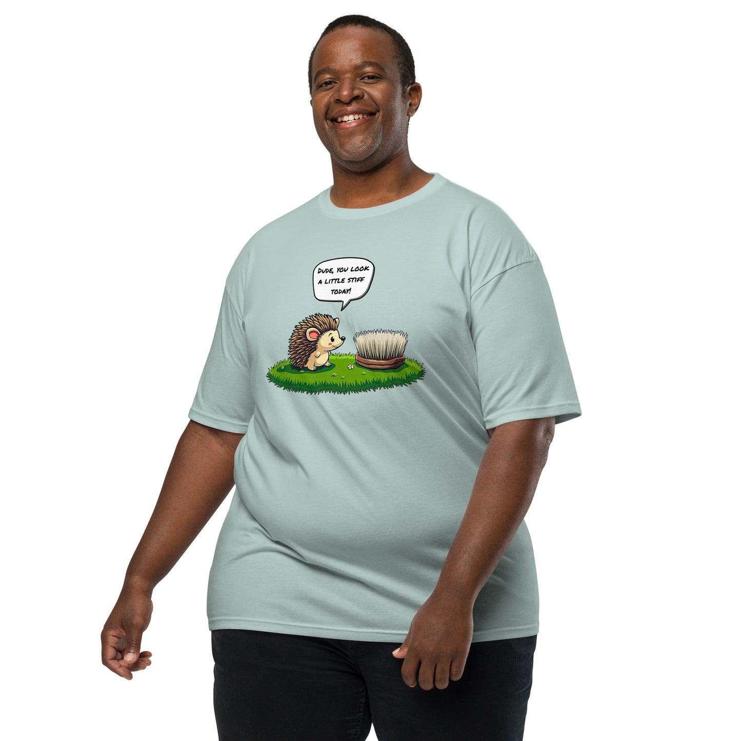 A smiling man wears a grabNade Men's Relaxed Heavyweight Tee in light blue, featuring a cartoon hedgehog with the text “Dude you look a little stiff today,” standing on green grass against a white background.