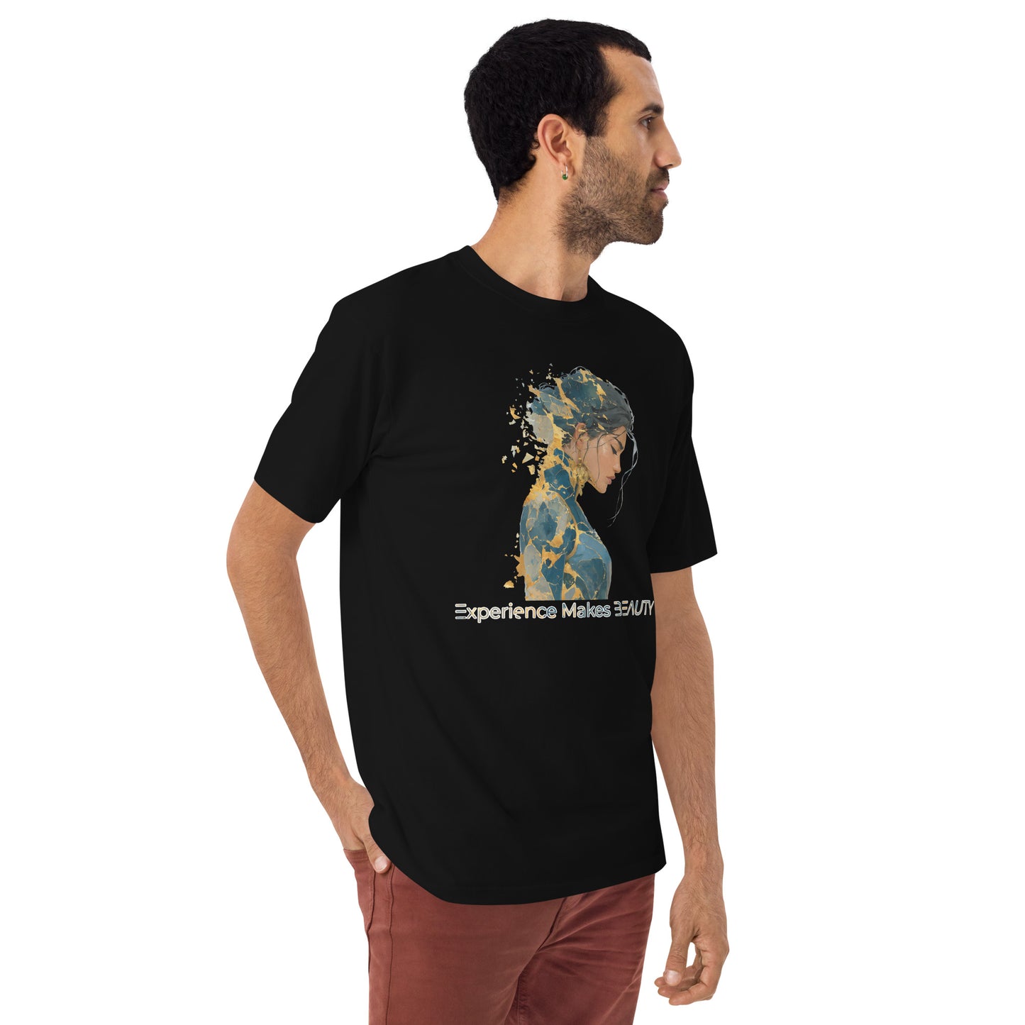 Product mockup. Man with short curly hair standing at an angle in a black heavyweight t-shirt and brick-colored pants, printed with a side-silhouette female portrait, gold paint splatter effect, and the words "Experience Makes BEAUTY."