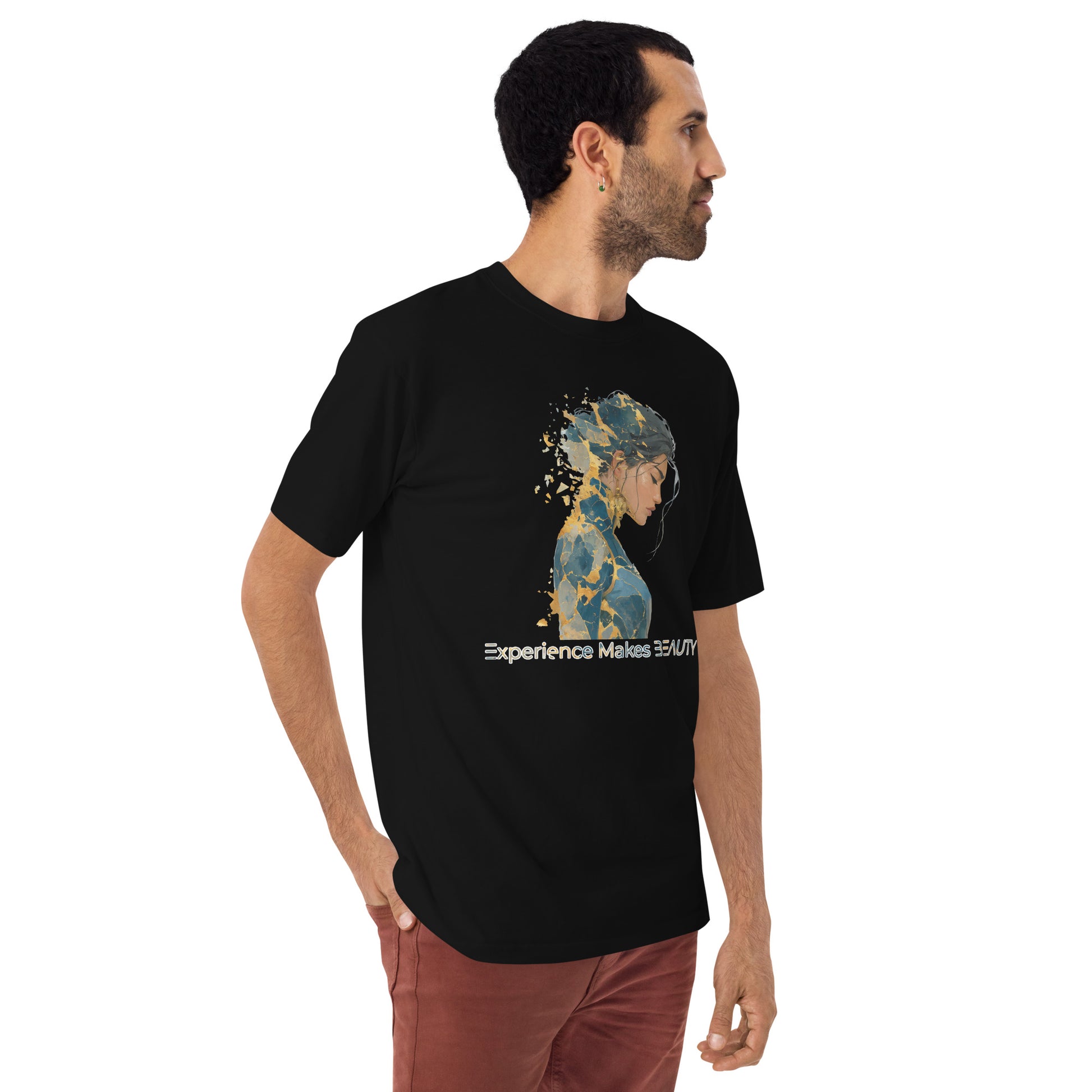 Product mockup. Man with short curly hair standing at an angle in a black heavyweight t-shirt and brick-colored pants, printed with a side-silhouette female portrait, gold paint splatter effect, and the words "Experience Makes BEAUTY."
