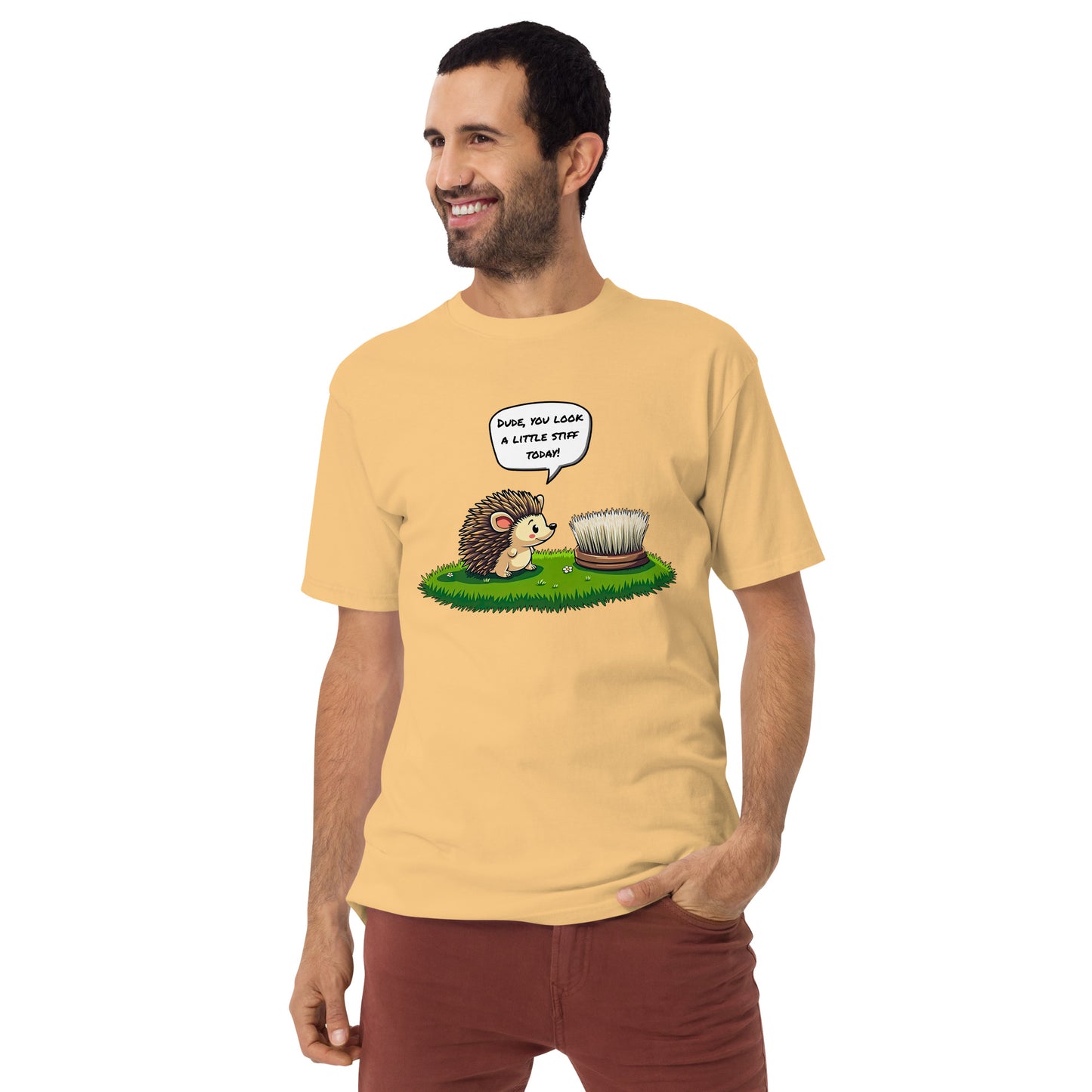 Product mockup. Man with short curly hair and an earring standing at an angle in a pale yellow t-shirt and brick-colored pants, with a cartoon hedgehog talking to a brush and the caption "Dude, you look a little stiff today!"