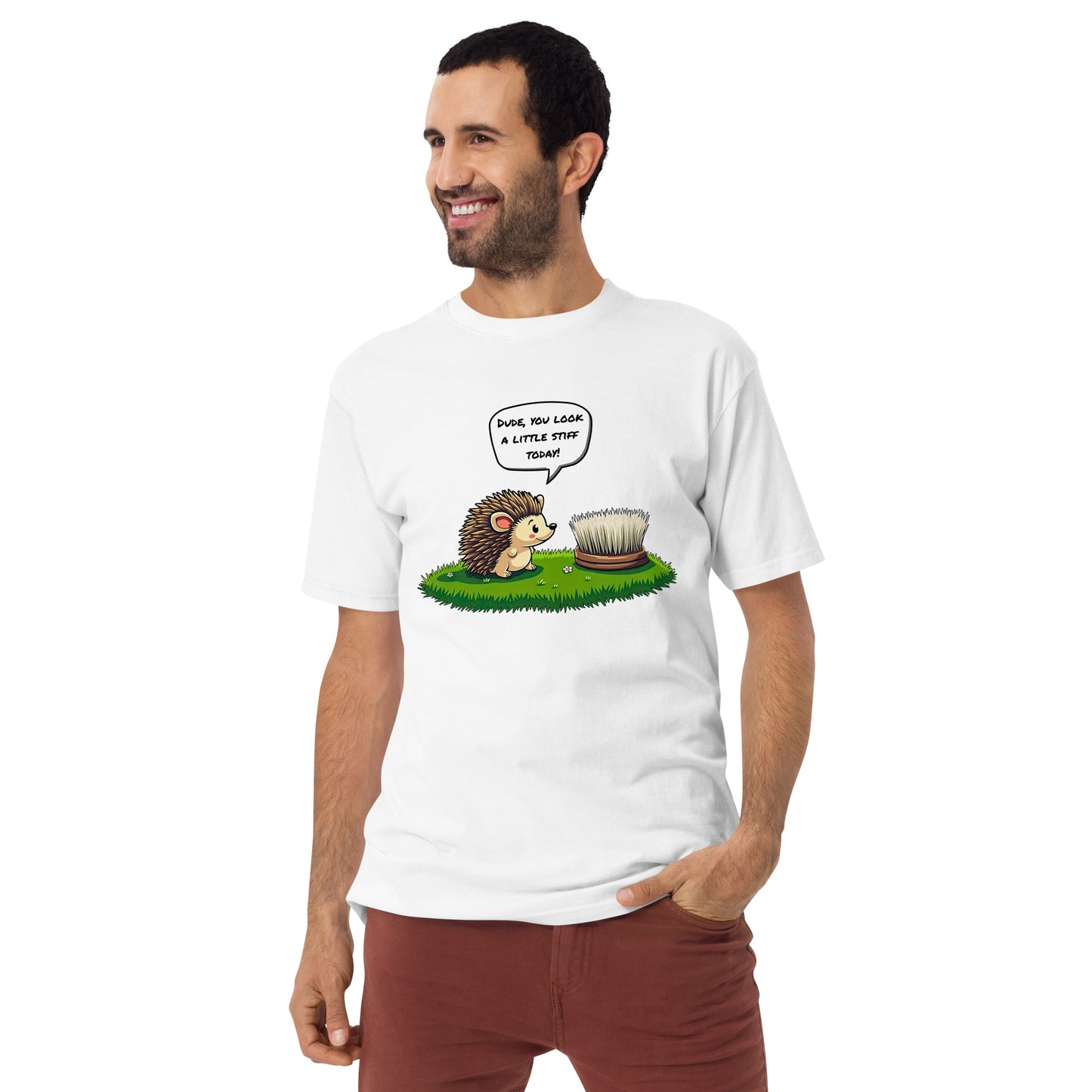 A smiling man with short dark hair and a beard, wearing the grabNade Men's Relaxed Heavyweight Tee in premium cotton. The shirt features a cartoon hedgehog telling a hairbrush, “Dude you look a little stiff today!” Both stand on green grass.