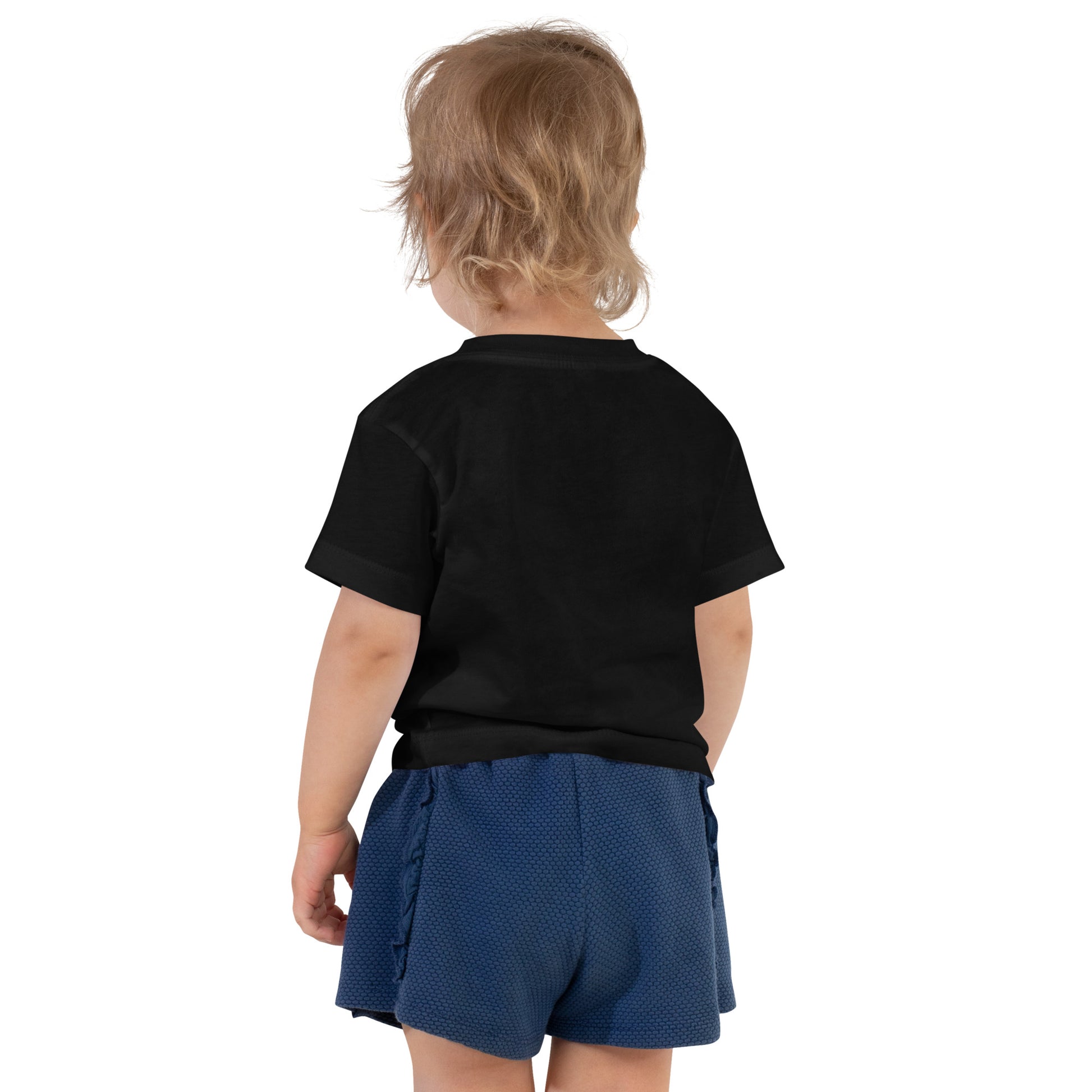 Product mockup. A toddler seen from behind, wearing a plain black short-sleeve t-shirt and textured navy blue shorts. The child's hair is medium length and slightly tousled, and no designs are visible on the back of the shirt.