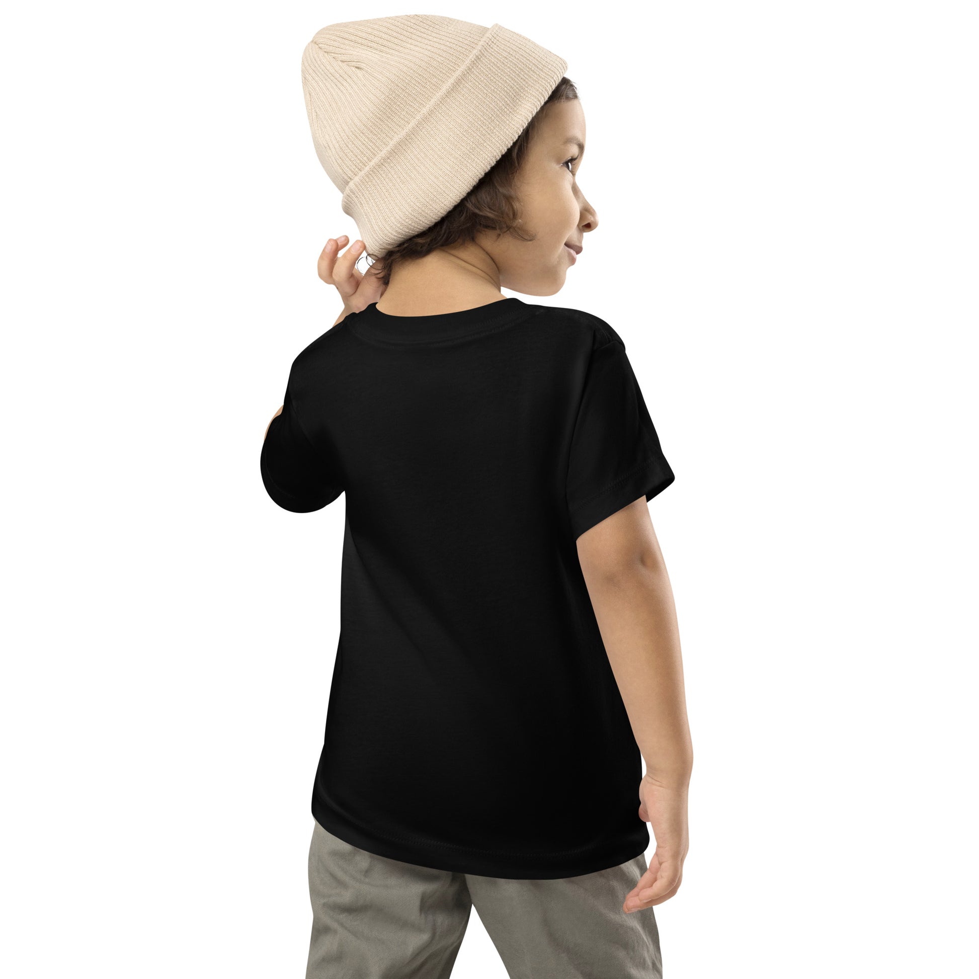Product mockup. A young child shown from behind, wearing a light beige knit beanie, a plain black t-shirt with no visible graphics on the back, and light-colored pants. The child is looking over their shoulder with one arm slightly raised
