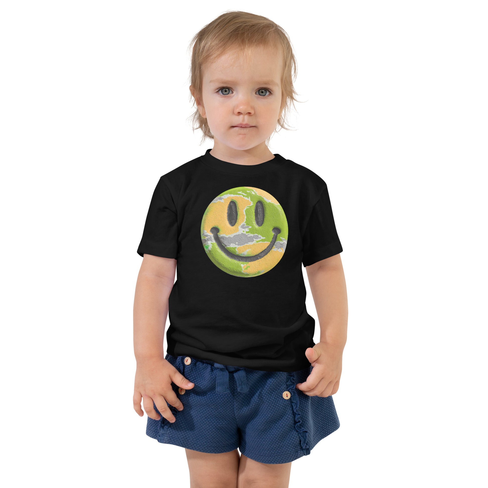 Product mockup. Toddler standing, wearing a black t-shirt with a blue, yellow, and green smiley face centered on the chest.