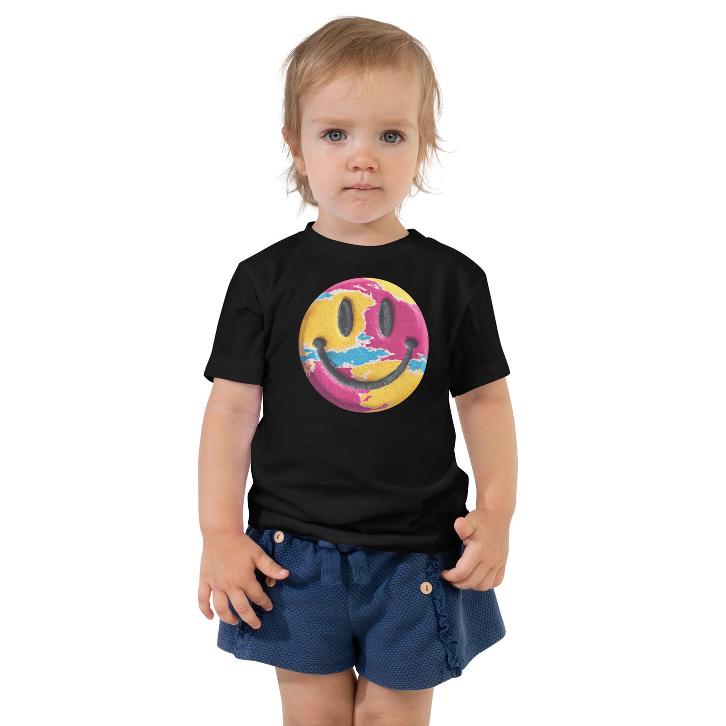 Product mockup. Toddler standing, wearing a black t-shirt with a blue, yellow, and green smiley face centered on the chest.