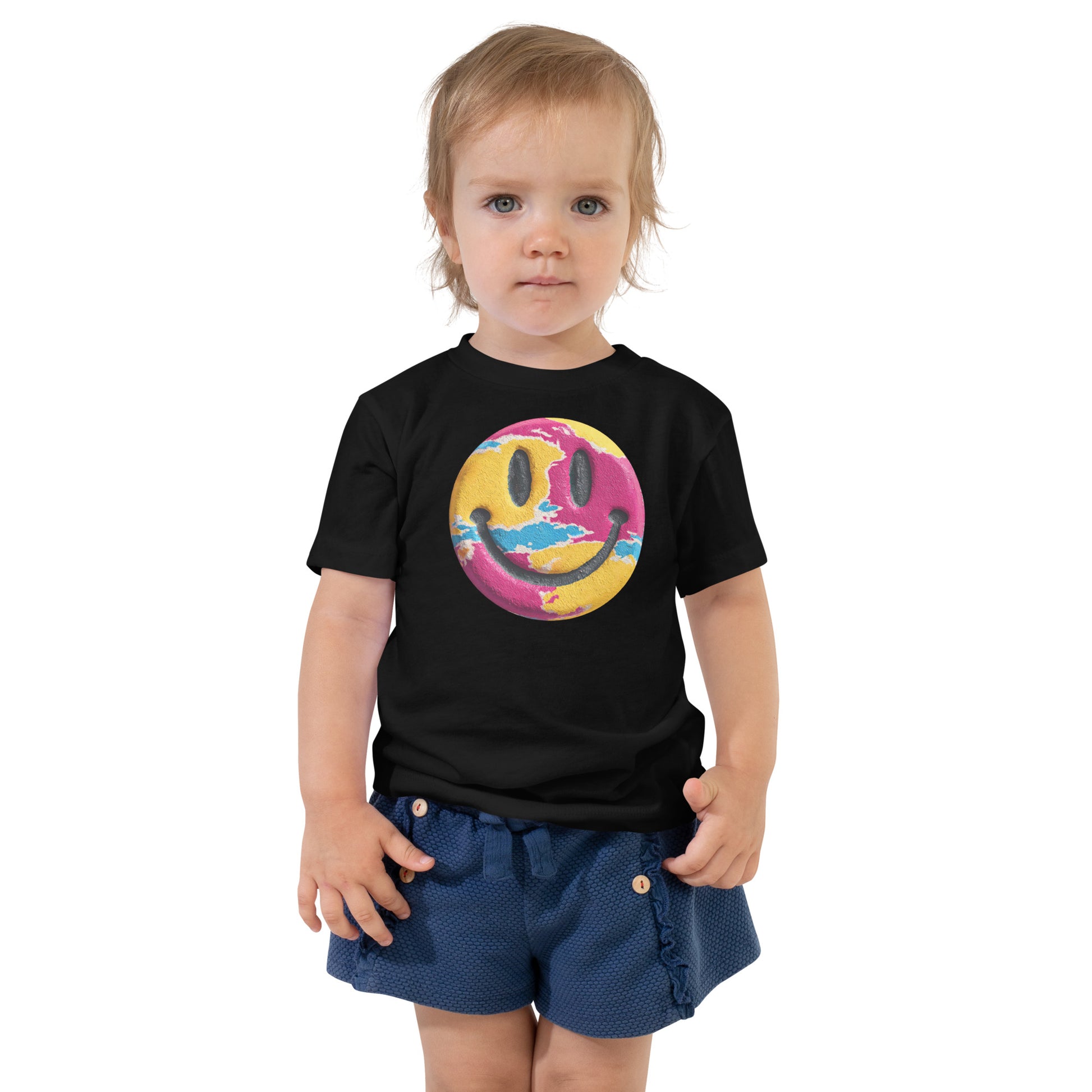 Product mockup. Toddler standing, wearing a black t-shirt with a blue, yellow, and green smiley face centered on the chest.