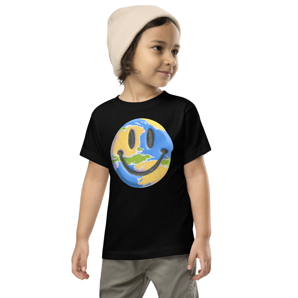 Product mockup. A young child wearing a cream knit beanie and a black t-shirt featuring a large central graphic of a smiley face designed to look like a world globe, with green continents, blue oceans, and black cartoon-style eyes and mouth.