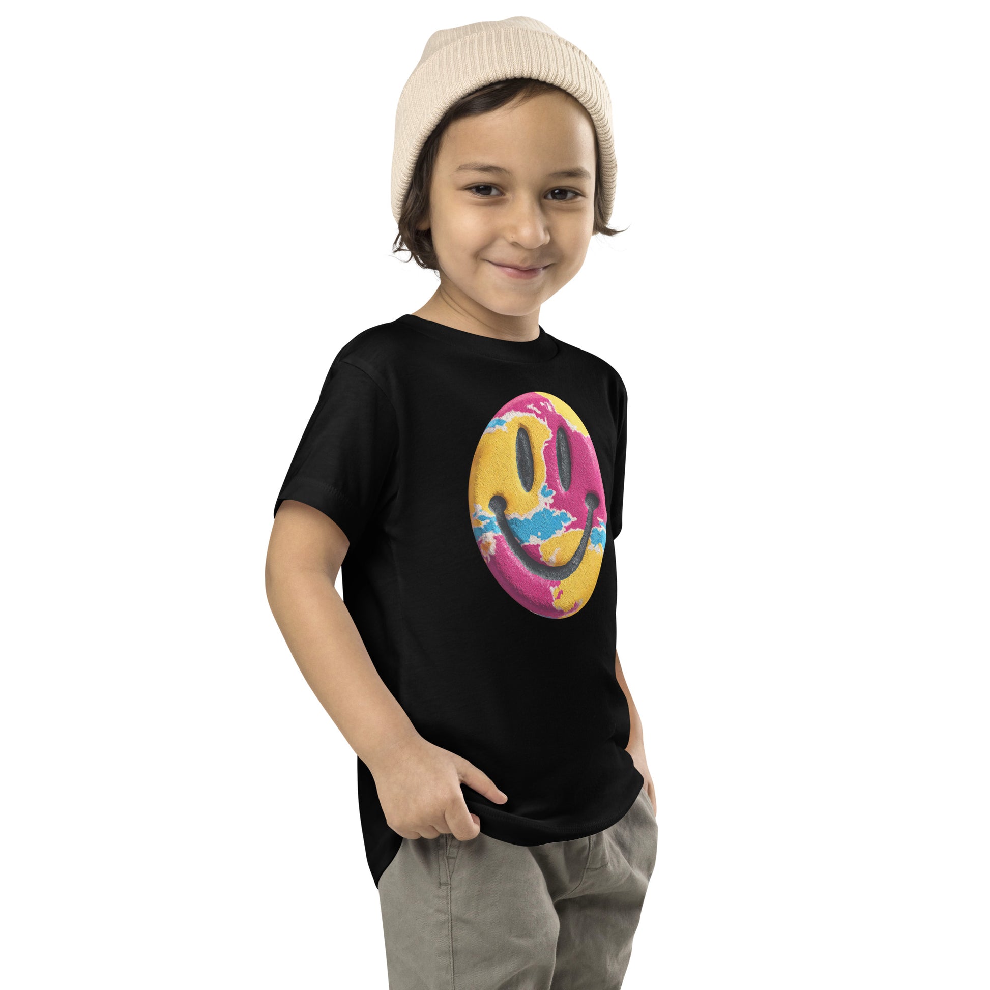 Product mockup. Child standing sideways, wearing a black t-shirt with a large pink, yellow, and blue smiley face icon, khaki pants, and a beige knit hat.
