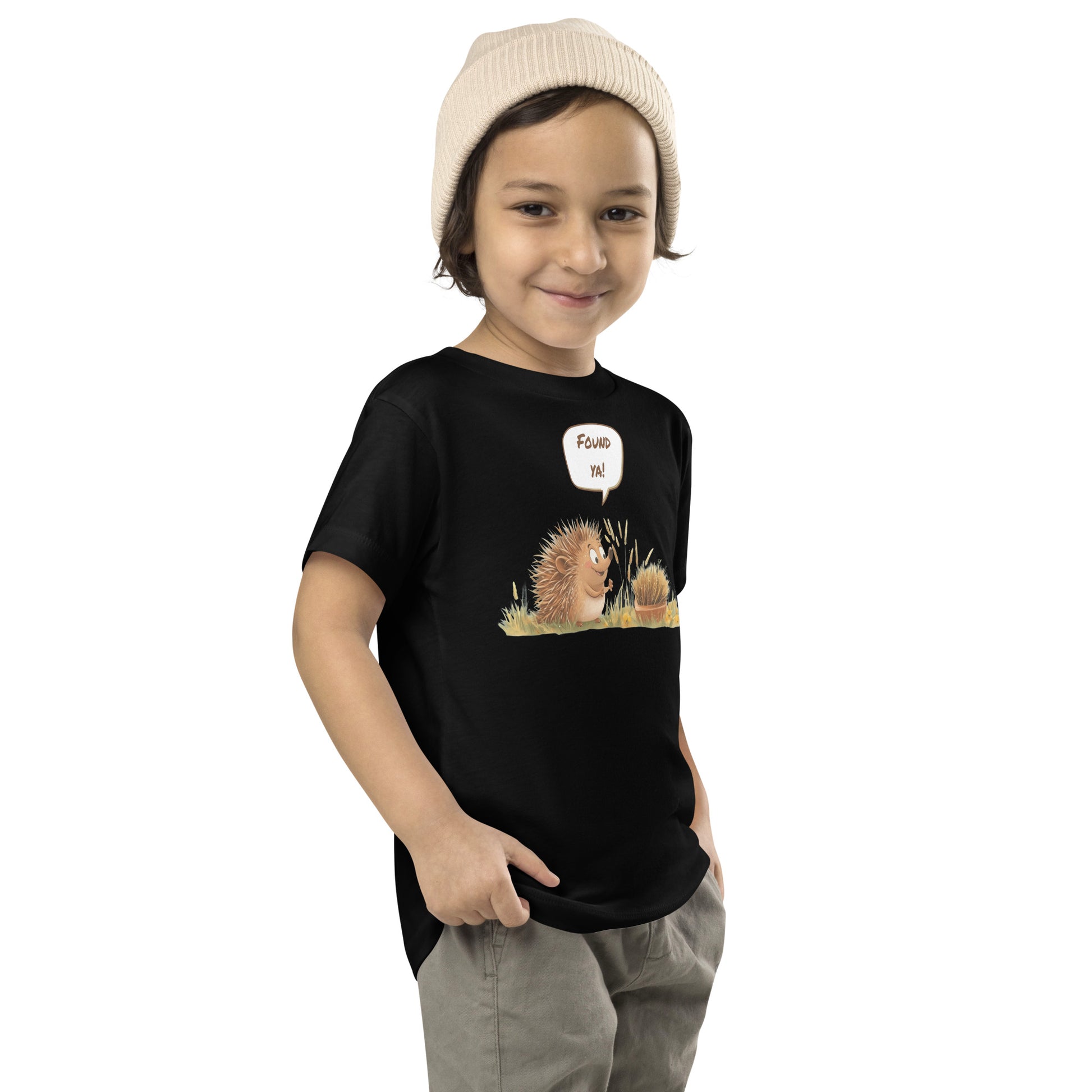 Product mockup. Child standing sideways, wearing a black t-shirt with a large pink, yellow, and blue smiley face icon, khaki pants, and a beige knit hat.