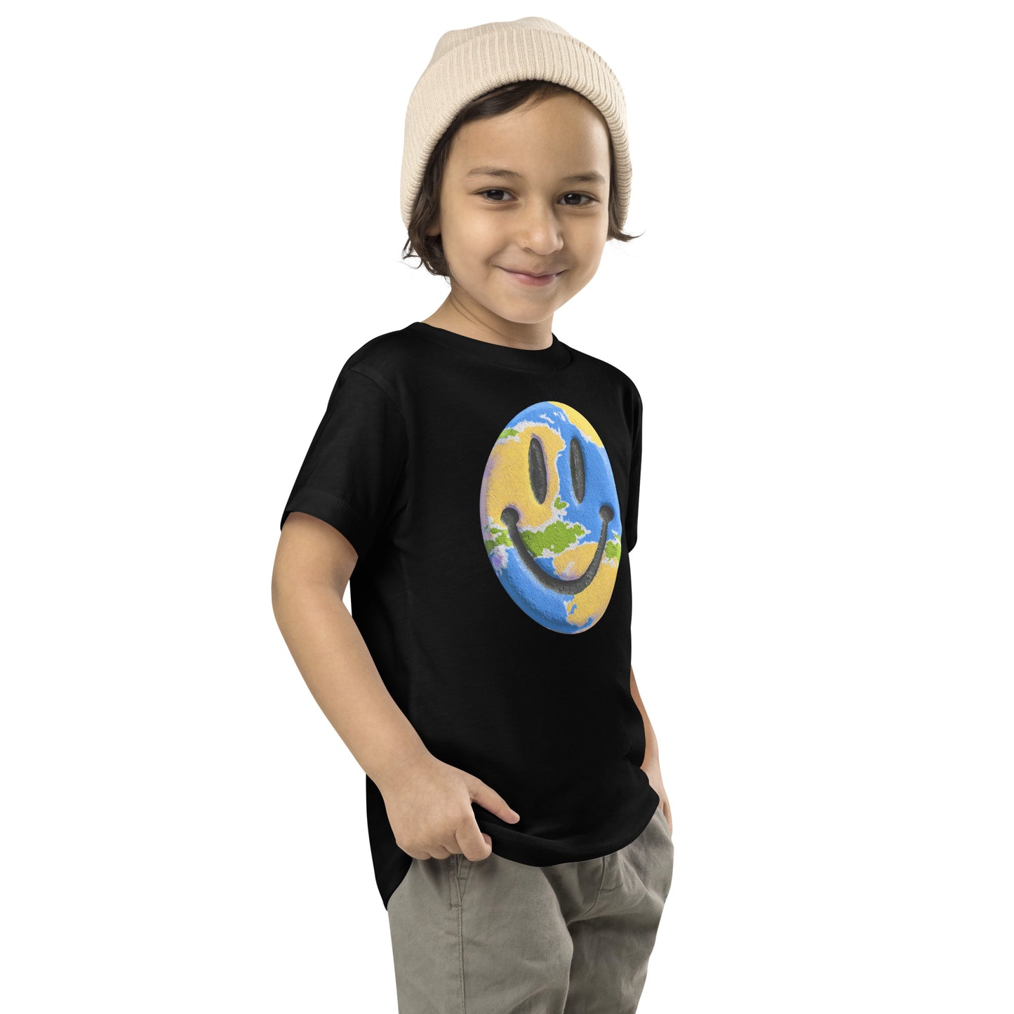 Product mockup. A young child wearing a cream knit beanie and a black t-shirt featuring a large central graphic of a smiley face designed to look like a world globe, with green continents, blue oceans, and black cartoon-style eyes and mouth.