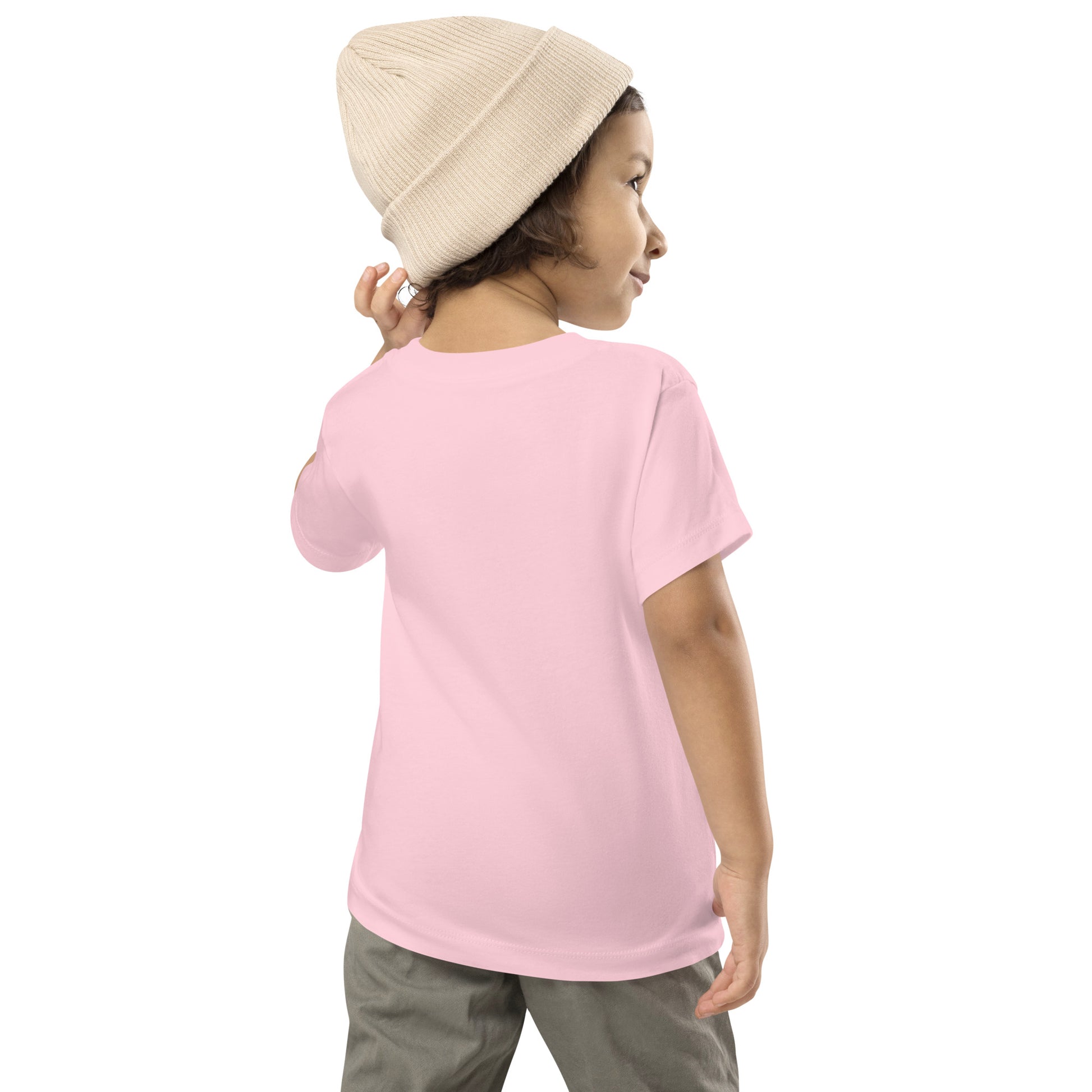 Product mockup. A young child shown from behind, wearing a light beige knit beanie, a plain light pink t-shirt with no visible graphics on the back, and light-colored pants. The child is looking over their shoulder with one arm slightly raised