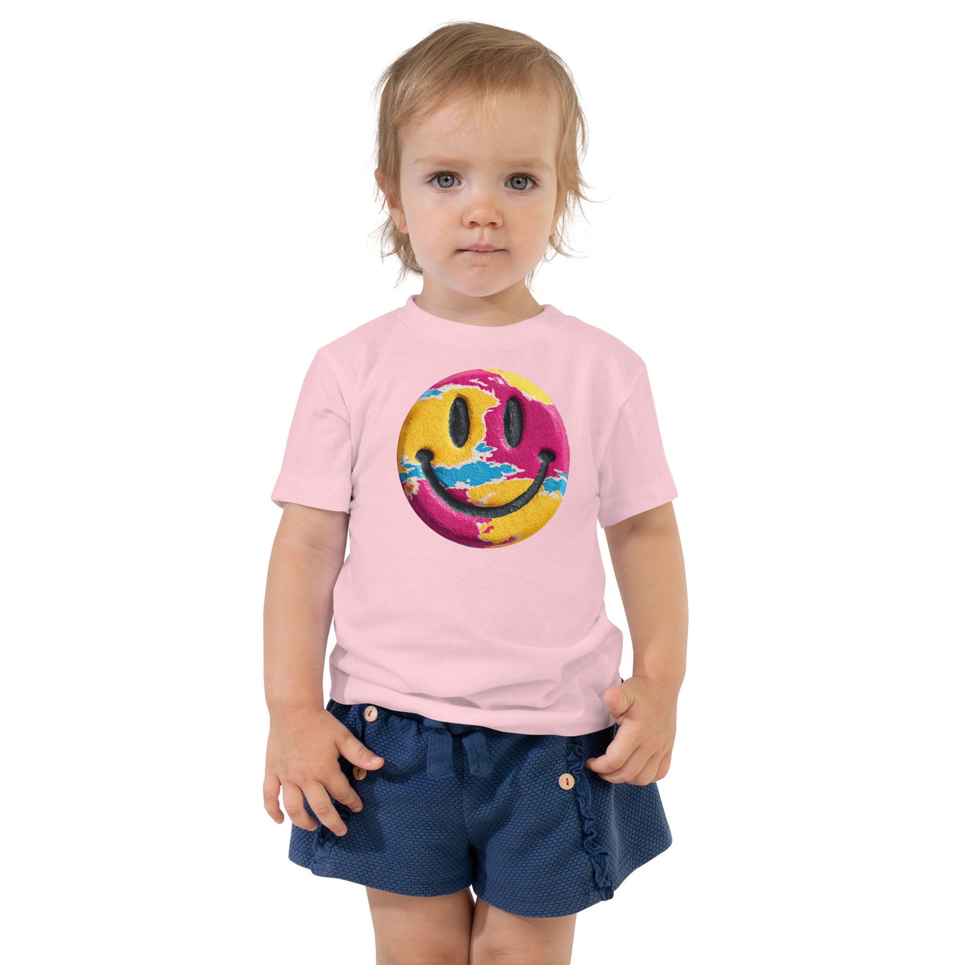 Product mockup. Toddler's light pink t-shirt laid flat, centered with a blue, yellow, and green smiley face graphic.