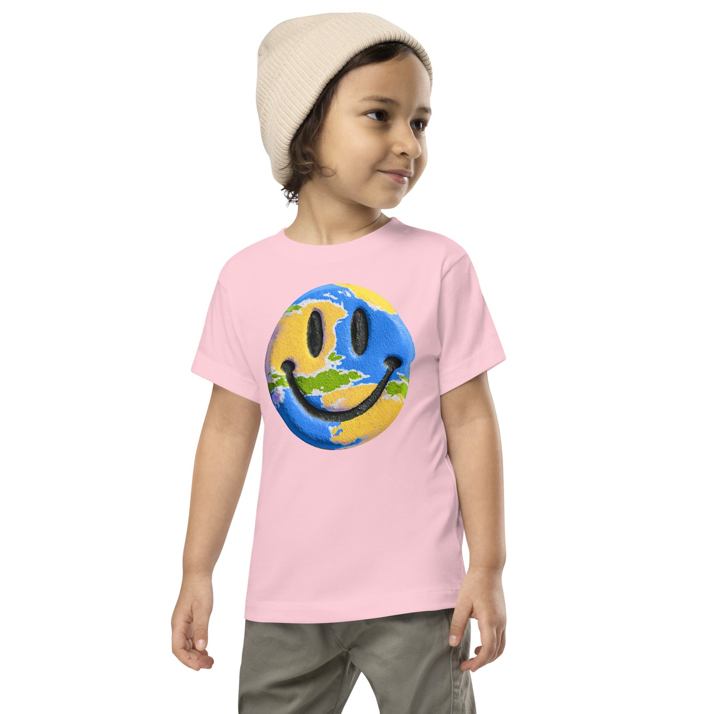 Product mockup. A young child wearing a cream knit beanie and a light pink t-shirt featuring a large central graphic of a smiley face designed to look like a world globe, with green continents, blue oceans, and black cartoon-style eyes and mouth.