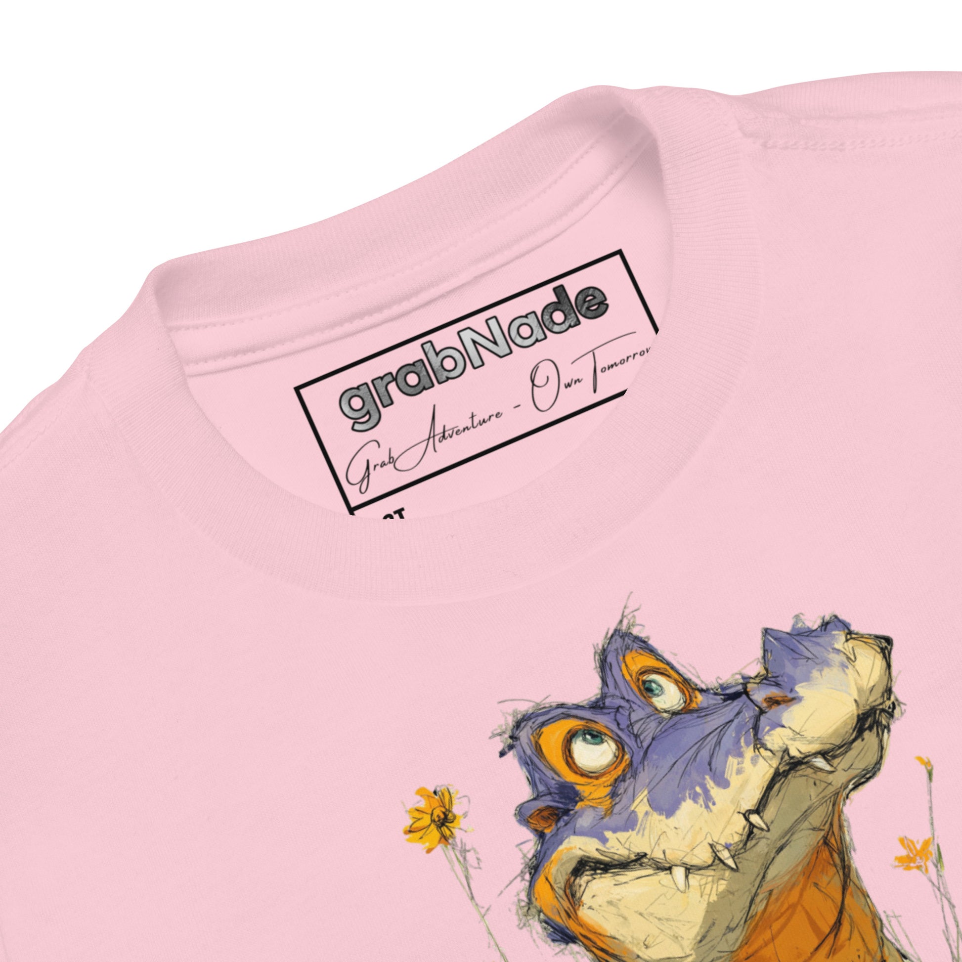 Product mockup. Close-up view of a light pink t-shirt collar showing the 'grabNade' brand label inside the neckline, with handwritten-style text beneath the brand name reading 'Grab Adventure - Own Tomorrow.' The shirt features part of a colorful cartoon illustration of a purple and orange dinosaur and yellow flowers visible below the collar.