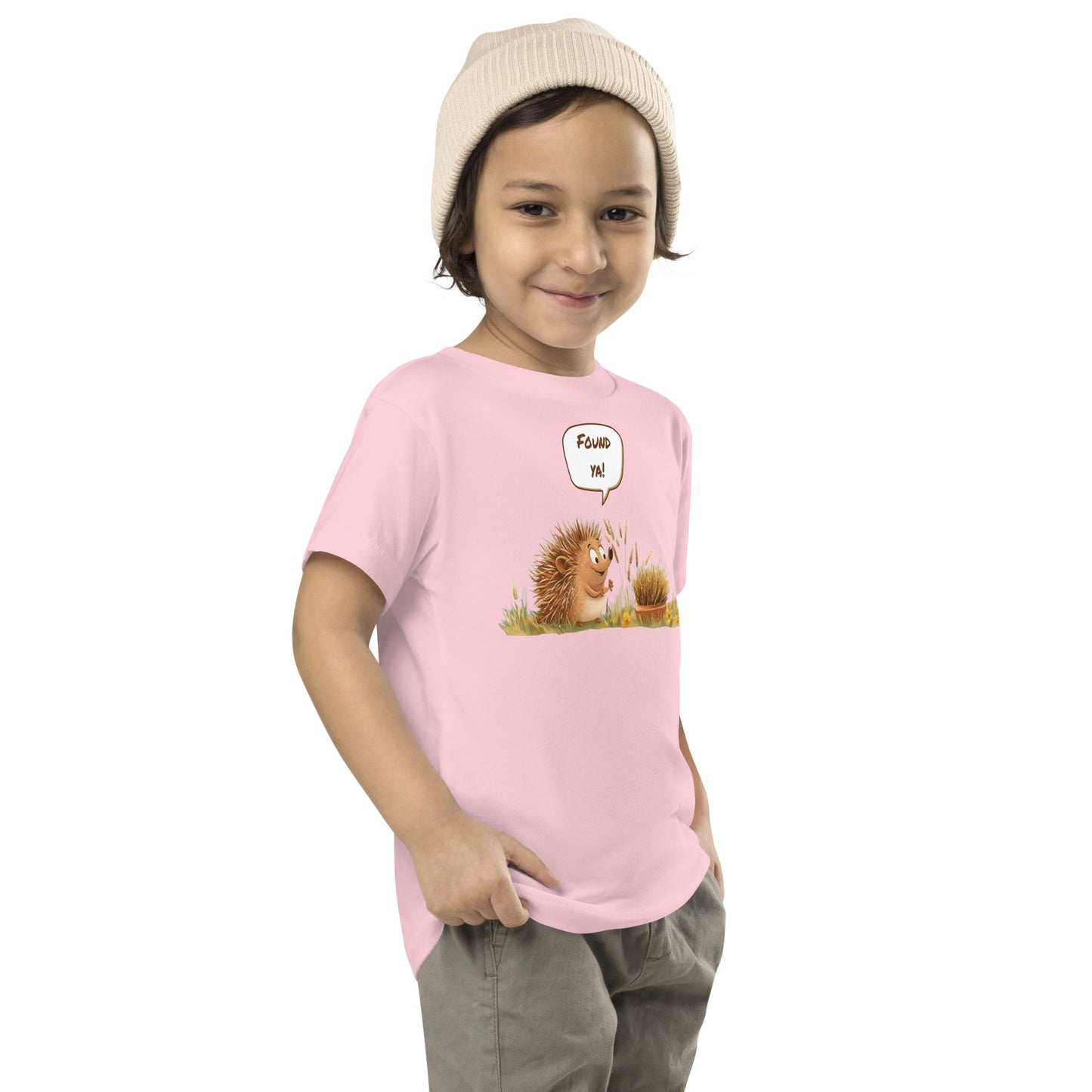 Product mockup. Child standing sideways, wearing a light pink t-shirt with a hedgehog cartoon saying "Found ya!", khaki pants, and beige hat.