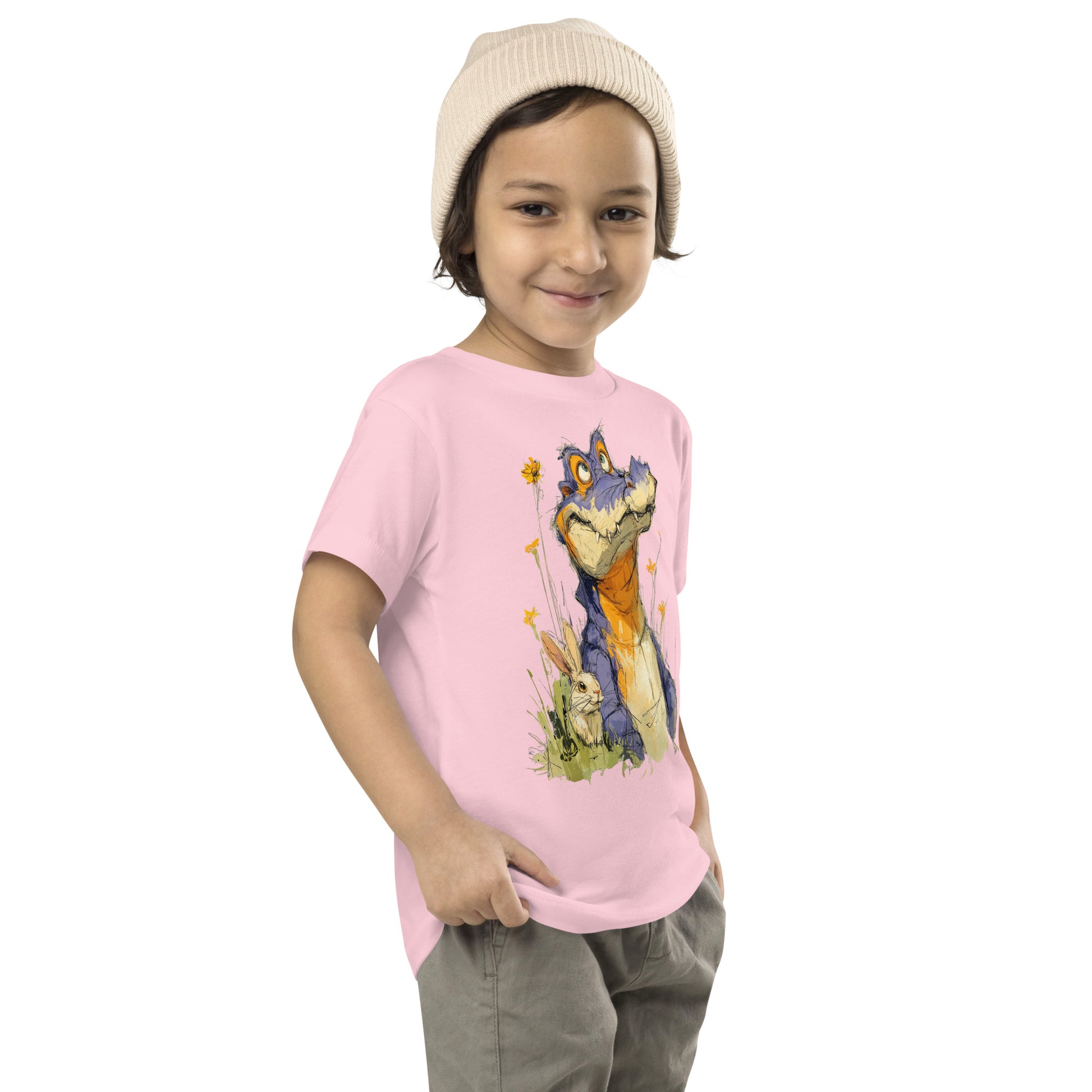 Product mockup. A young child wearing a light beige knit beanie and a light pink t-shirt with a large, whimsical illustration of a friendly purple and orange cartoon dinosaur surrounded by yellow wildflowers, with a small rabbit nestled in the grass near its feet.