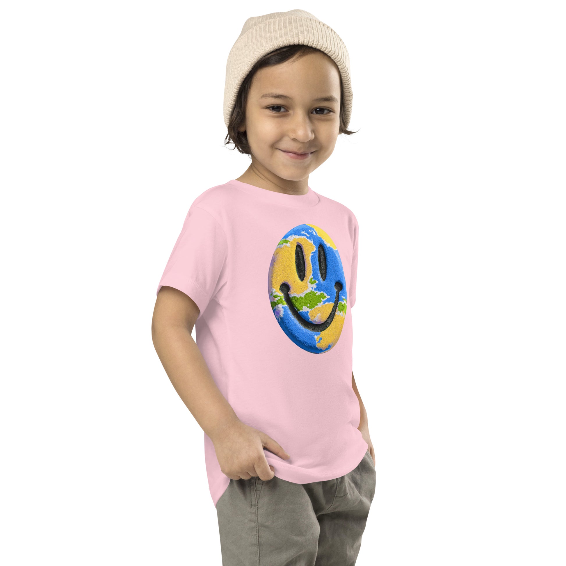 Product mockup. A young child wearing a cream knit beanie and a light pink t-shirt featuring a large central graphic of a smiley face designed to look like a world globe, with green continents, blue oceans, and black cartoon-style eyes and mouth.