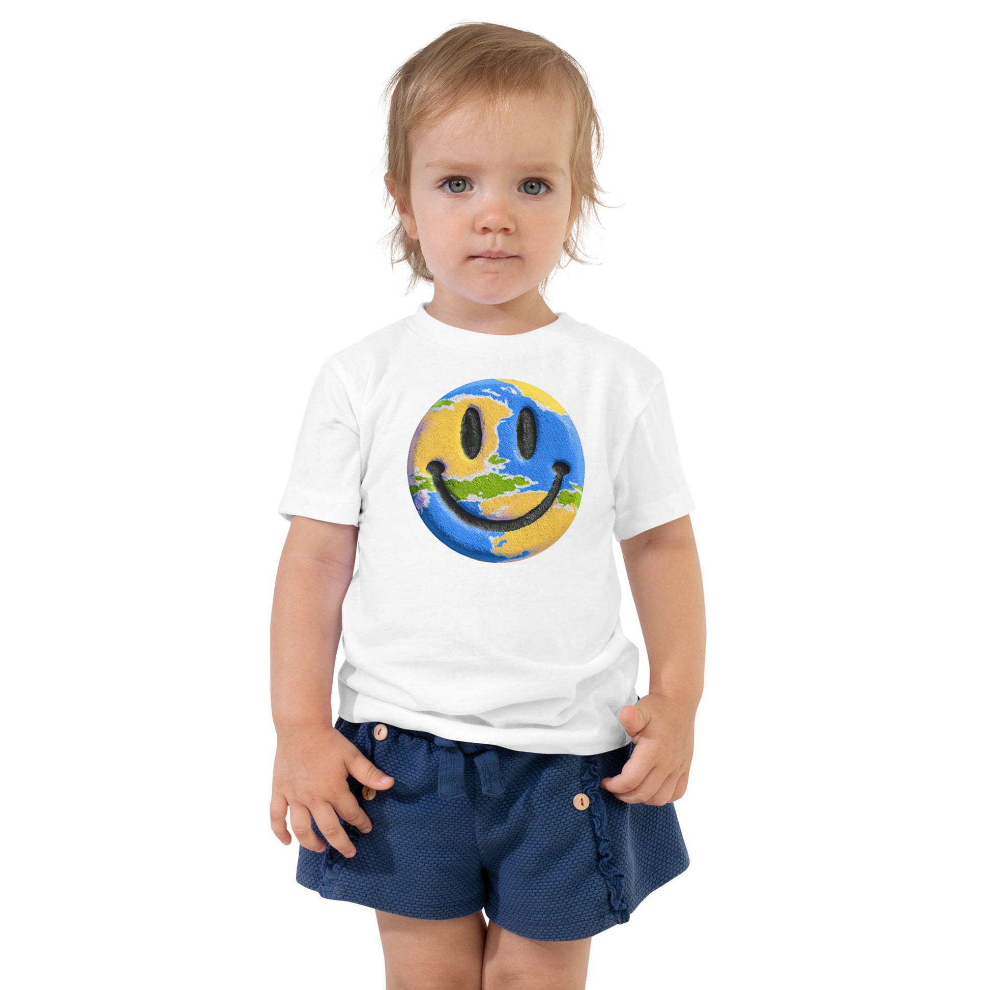 Product mockup. Toddler standing, wearing a white t-shirt with a large blue, yellow, and green smiley face icon, and blue jeans.