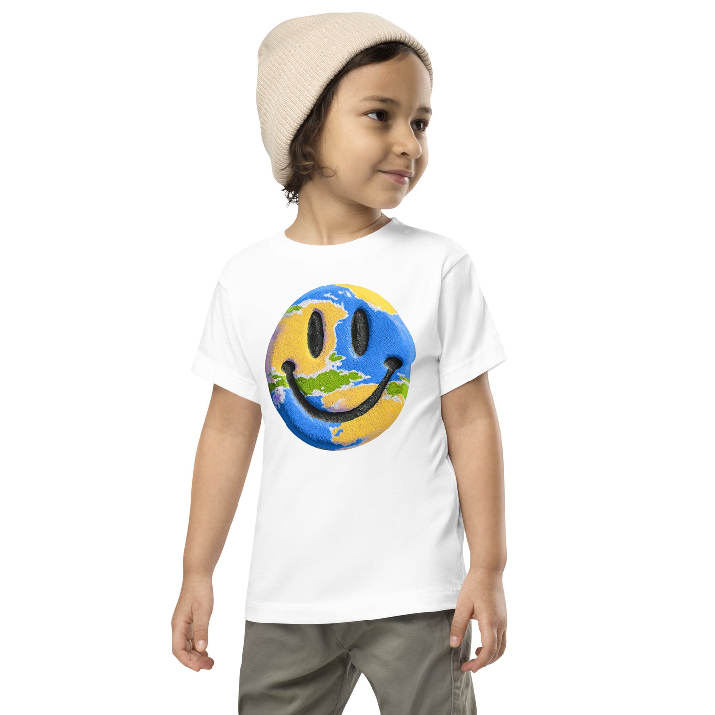 Product mockup. A young child wearing a cream knit beanie and a white t-shirt featuring a large central graphic of a smiley face designed to look like a world globe, with green continents, blue oceans, and black cartoon-style eyes and mouth.