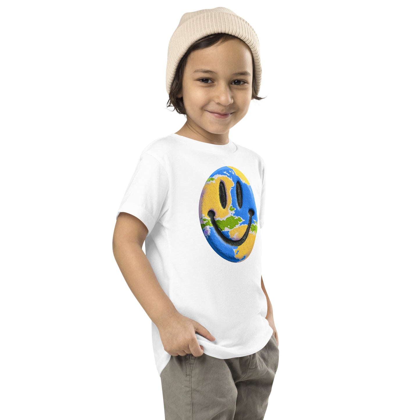 Product mockup. A young child wearing a cream knit beanie and a white t-shirt featuring a large central graphic of a smiley face designed to look like a world globe, with green continents, blue oceans, and black cartoon-style eyes and mouth.