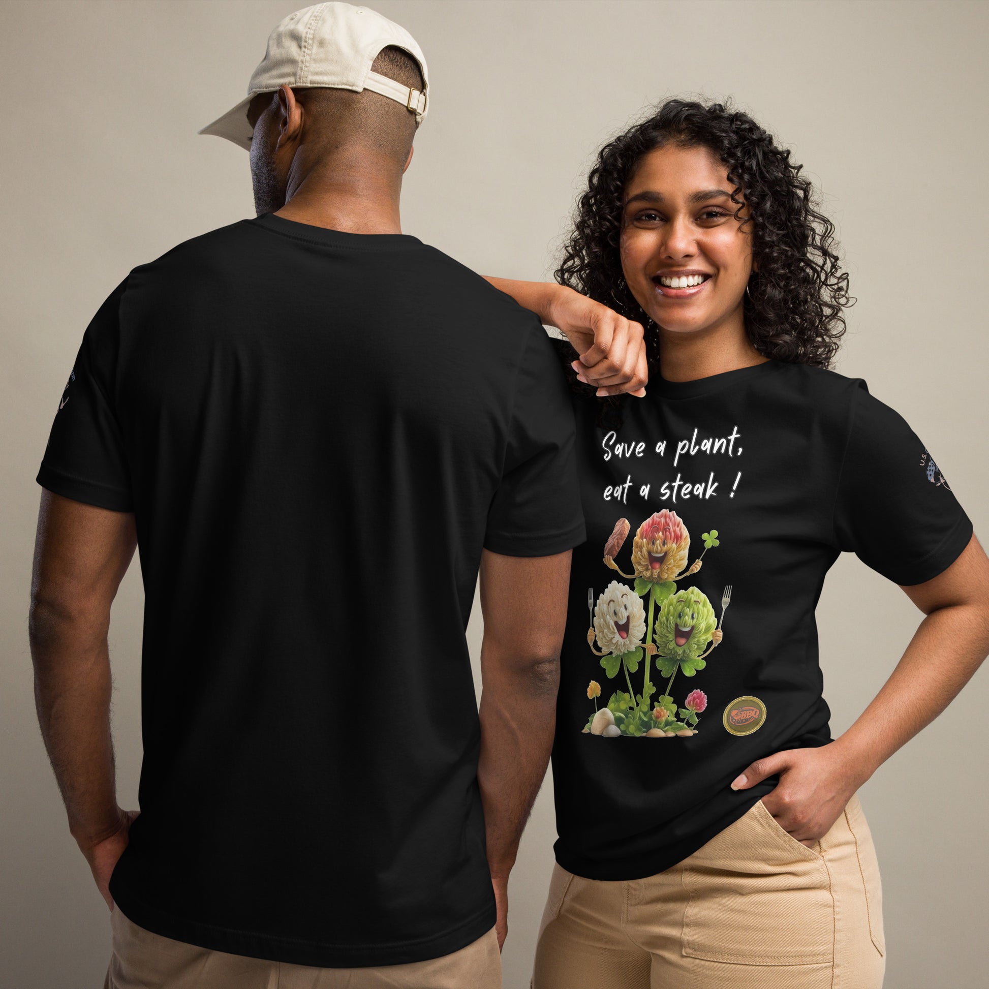 Product mockup. A man and woman stand together; the woman wears a black t-shirt with the phrase "Save a plant, eat a steak!" above a flower illustration holding a steak and clover, while the man displays the plain black back of his t-shirt.