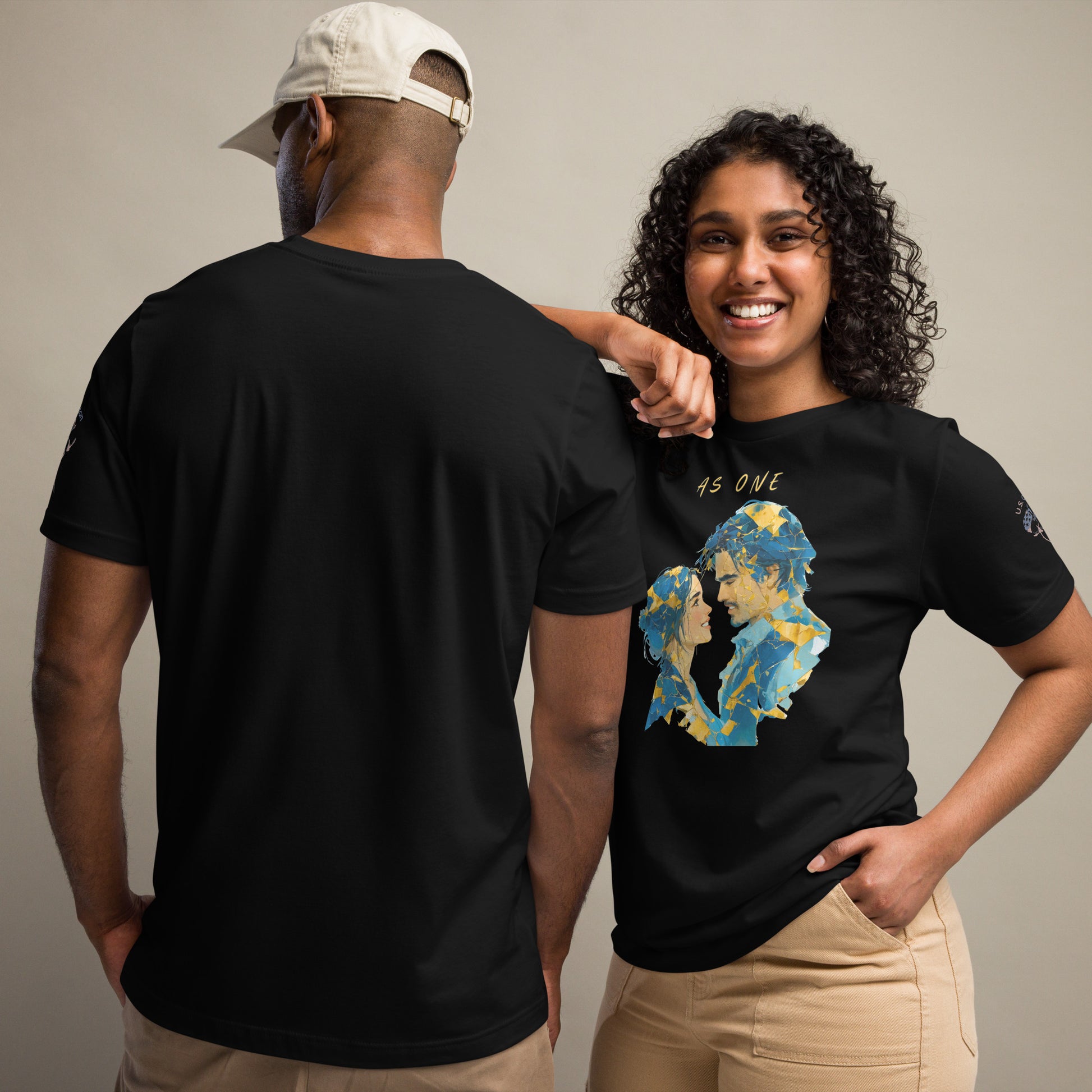 Product mockup. A man and woman stand together; the woman wears a black t-shirt with an abstract blue and yellow graphic and the phrase "AS ONE," while the man shows the back of a plain black t-shirt.