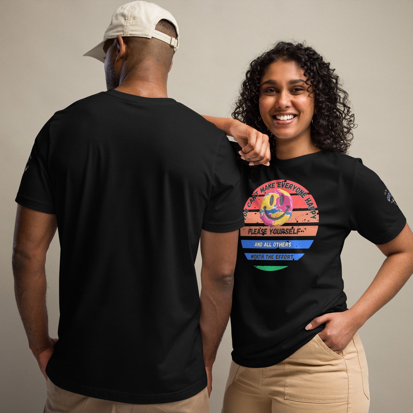 Product mockup. A man and woman stand together; the woman wears a black t-shirt with a striped circle, smiley face, and motivational text, while the man shows the back of a plain black t-shirt.