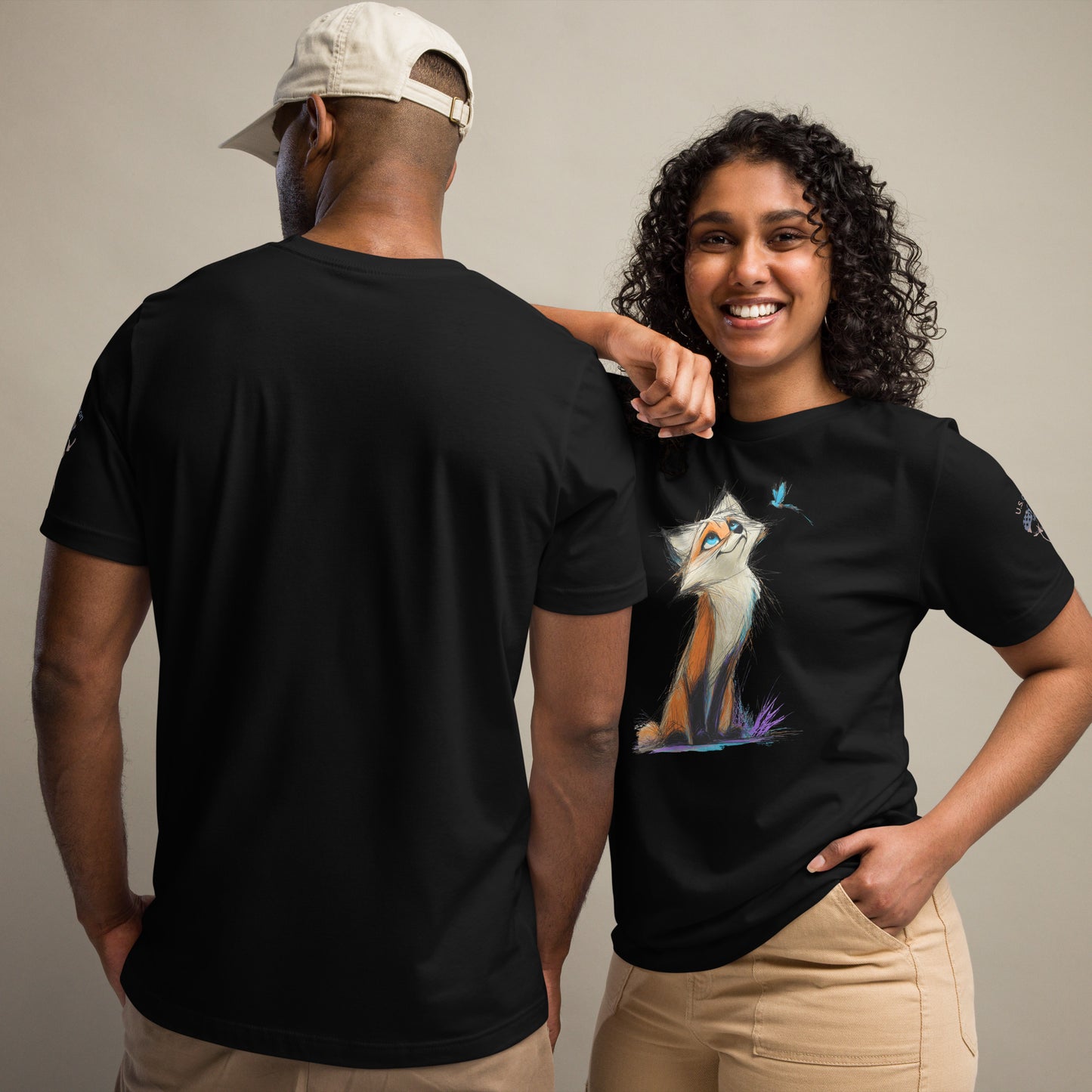 Product mockup. A man and woman stand together; the woman wears a black t-shirt with an illustrated orange and white fox sitting and looking up at a butterfly, while the man displays the back of a plain black t-shirt.