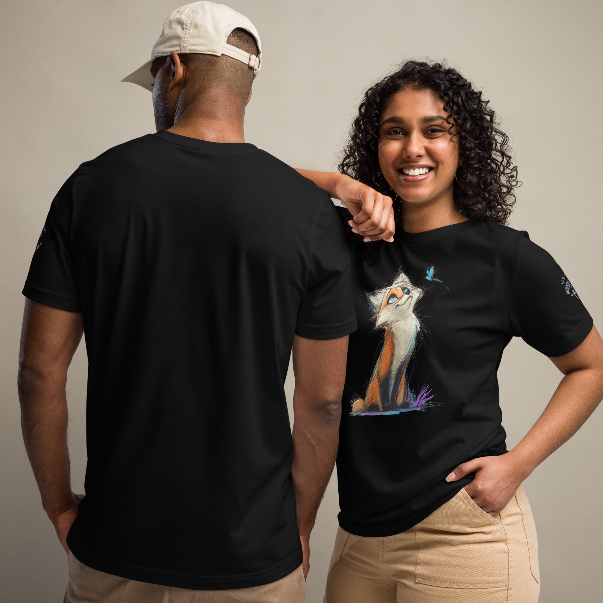 Product mockup. A man and woman stand together; the woman wears a black t-shirt with an illustrated orange and white fox sitting and looking up at a butterfly, while the man displays the back of a plain black t-shirt.