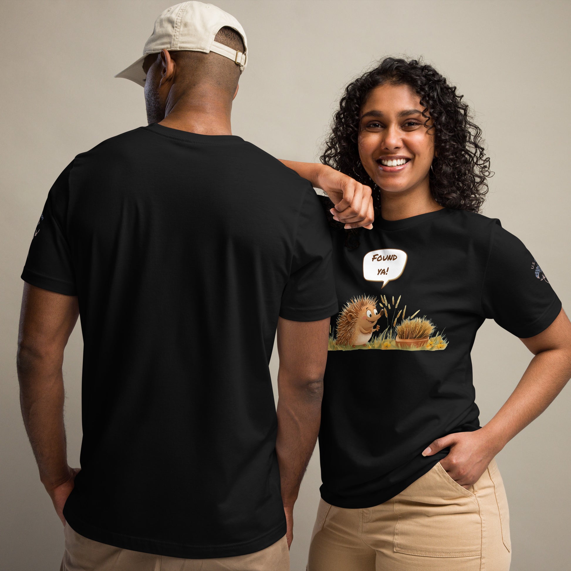 Product mockup. A man and woman stand together; the woman wears a black t-shirt with a cartoon hedgehog saying "Found ya!" to another in the grass, while the man shows the back of a plain black t-shirt.