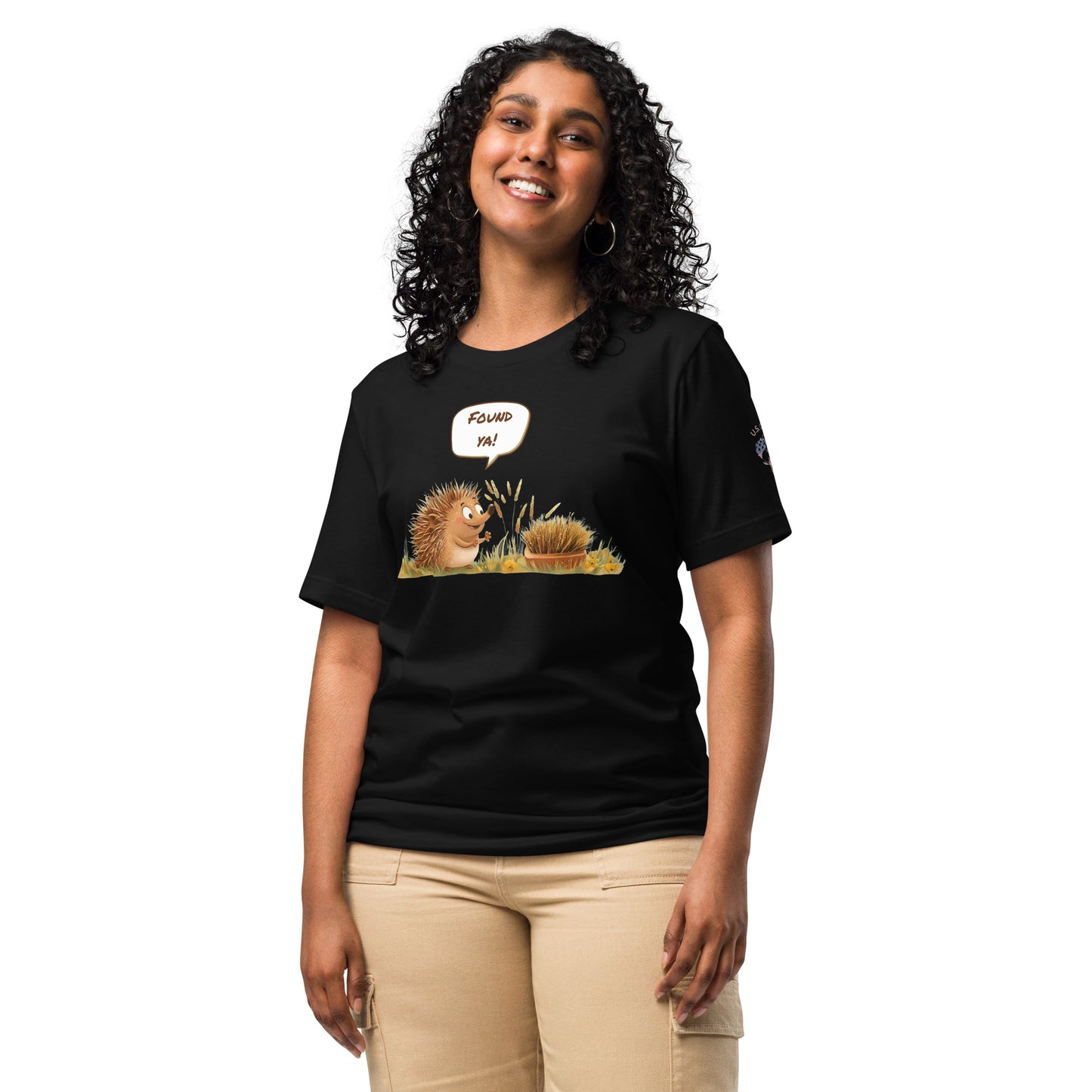 Product mockup. A woman stands wearing a black t-shirt with a cheerful hedgehog and grass design; a speech bubble says "Found ya!".