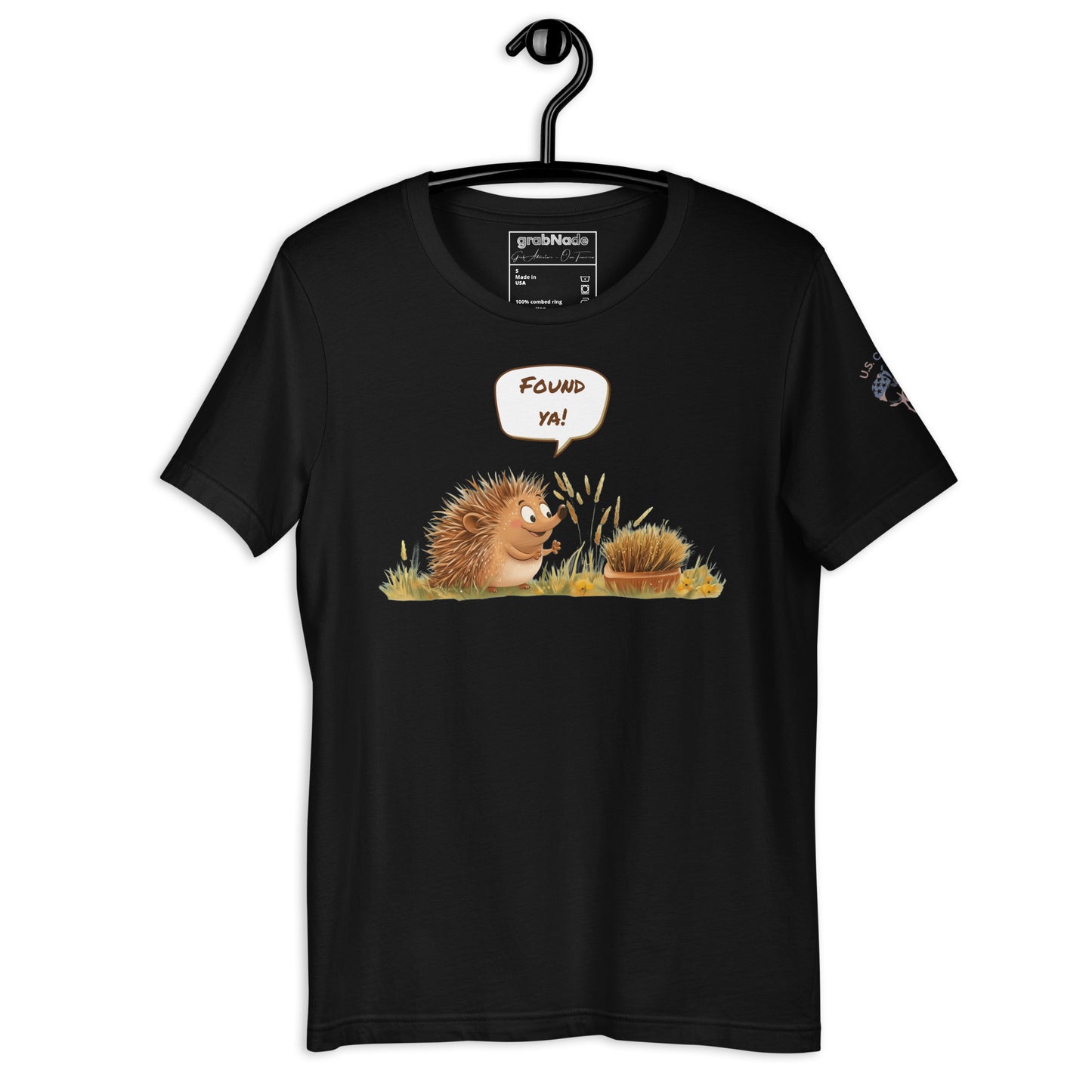 Product mockup. Black t-shirt on a hanger featuring a cartoon-style hedgehog saying "Found ya!" to another hidden hedgehog in grass.
