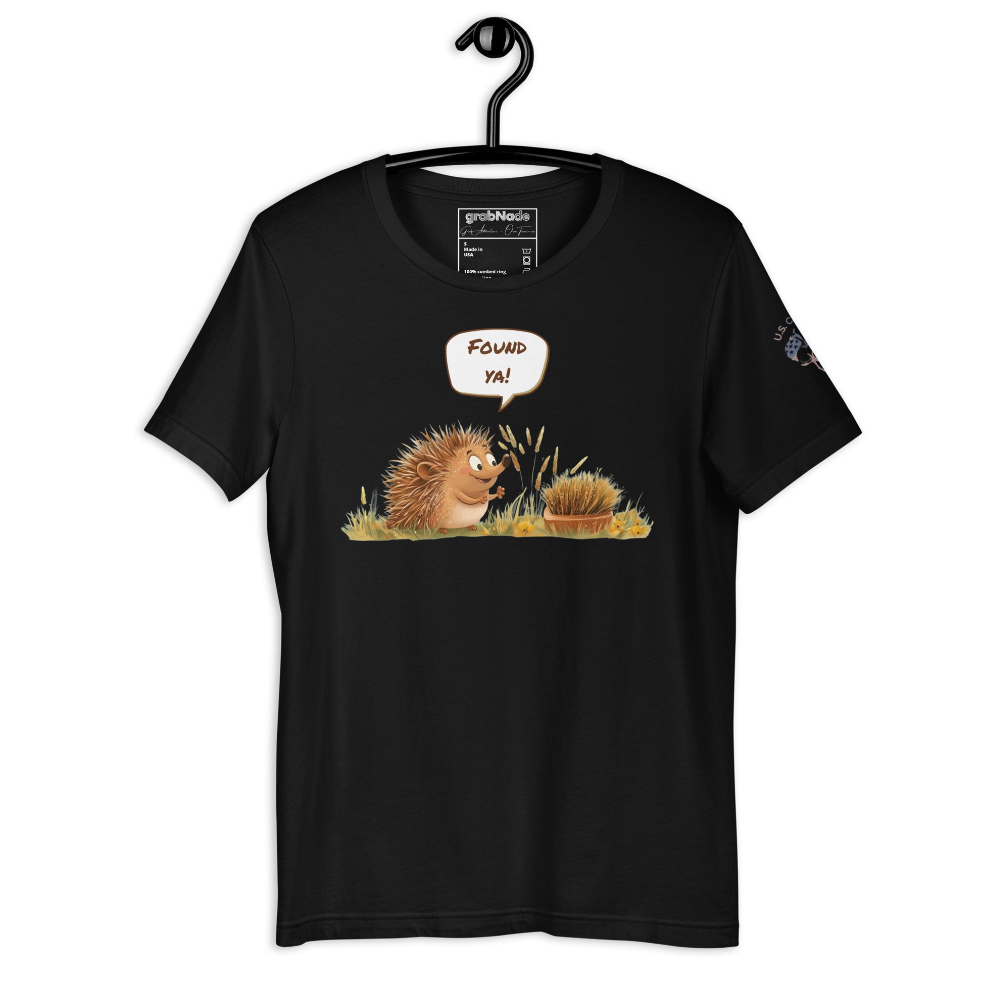 Product mockup. Black t-shirt on a hanger featuring a cartoon-style hedgehog saying "Found ya!" to another hidden hedgehog in grass.