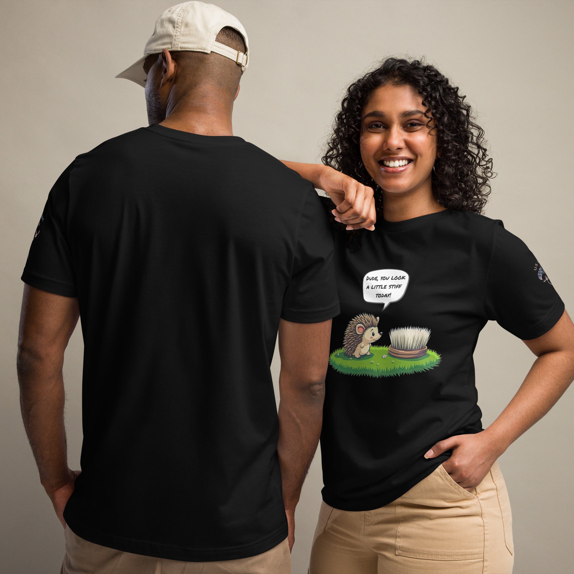 Product mockup. Man and woman in black t-shirts; woman in beige pants, shirt printed with a cute cartoon hedgehog looking at a stiff brush, speech bubble: “DUDE, YOU LOOK A LITTLE STIFF TODAY!” Man’s back view.