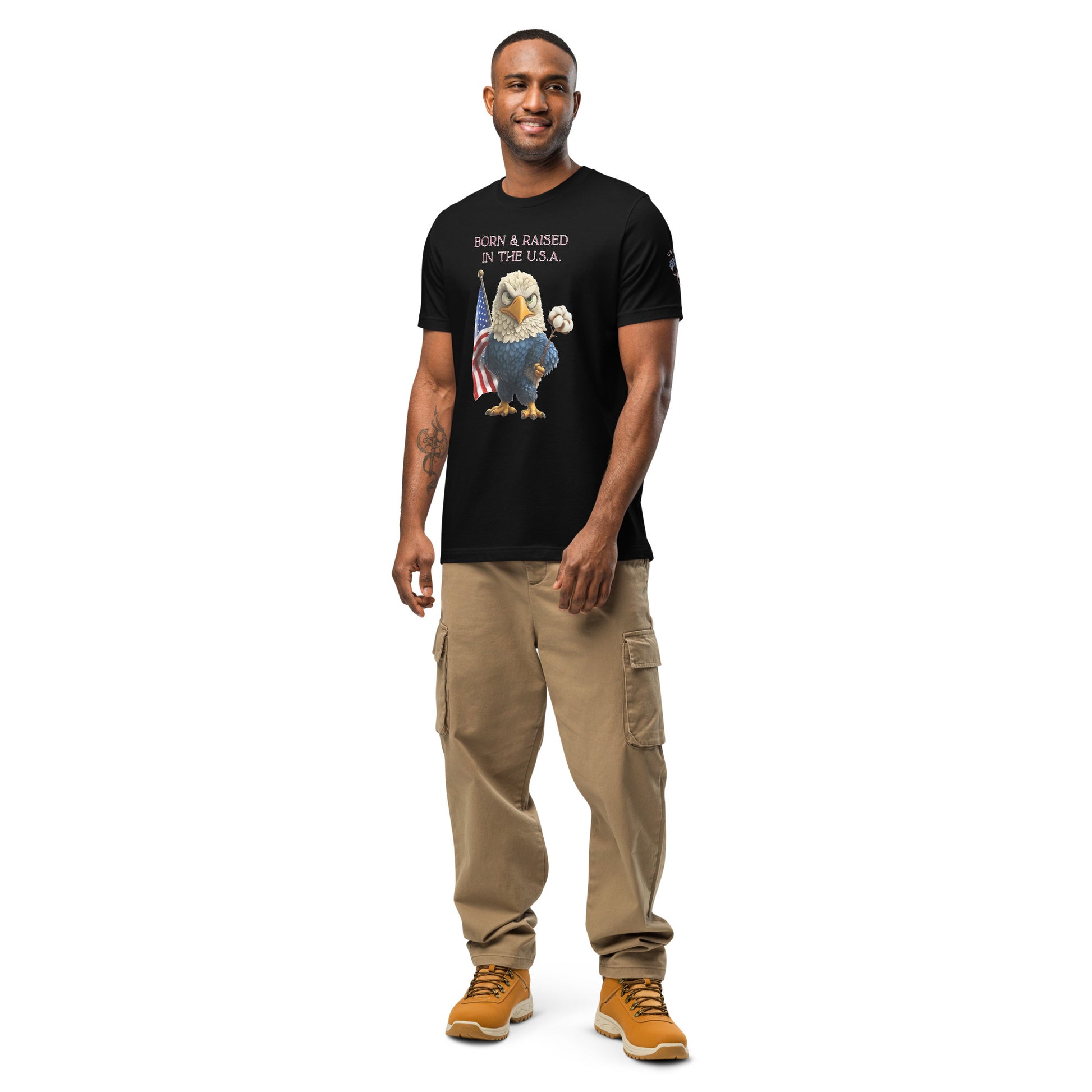 Product mockup. Person standing wearing black T-shirt displaying stylized eagle holding American flag and cotton stem with 'Born & Raised in the U.S.A.' text, khaki cargo pants, and tan boots.