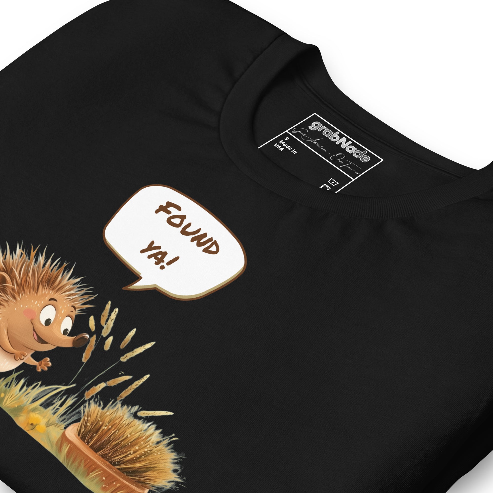 Product mockup. Close-up of a black t-shirt showing a cartoon hedgehog saying "FOUND YA!" to a round scrubbing brush on grass.