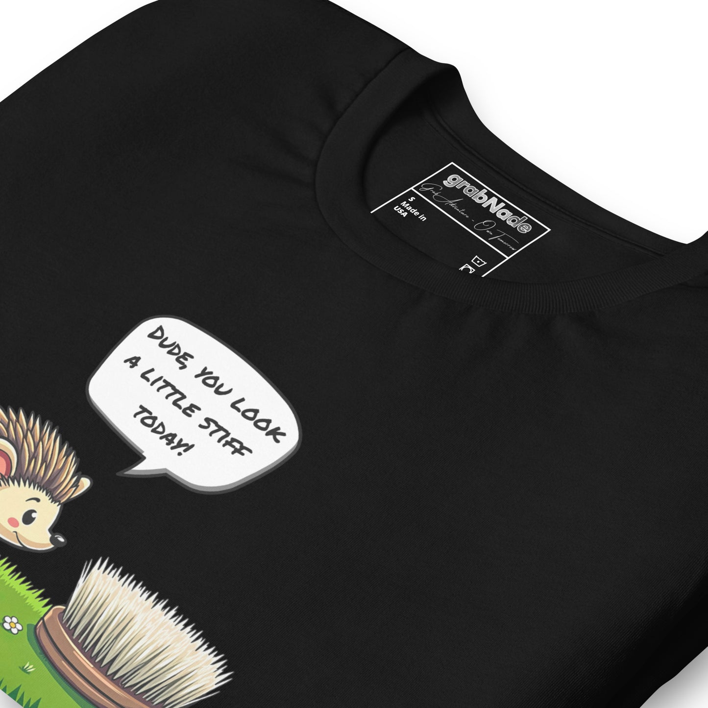 Product mockup. Close-up of a black t-shirt featuring a cartoon hedgehog talking to a scrubbing brush with the phrase "Dude, you look a little stiff today!" in a speech bubble.