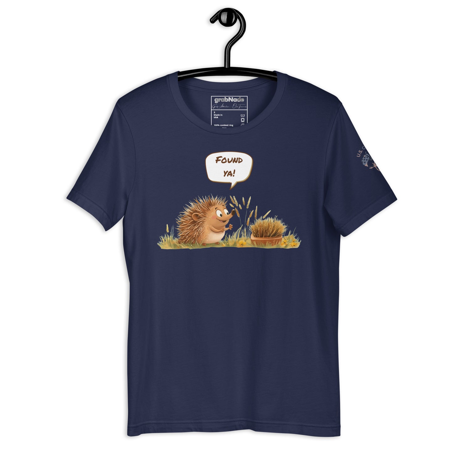 Product mockup. Navy blue t-shirt on a hanger featuring a cartoon-style hedgehog saying "Found ya!" to another hidden hedgehog in grass.