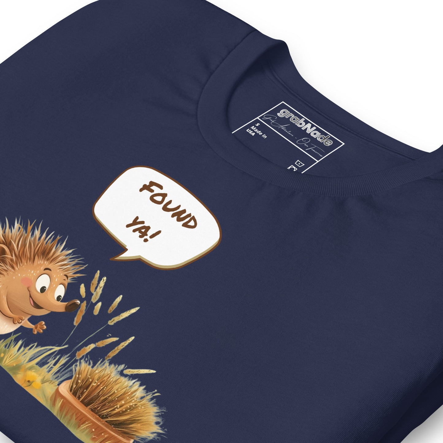 Product mockup. Close-up of navy blue T-shirt showing a cartoon hedgehog with a speech bubble "FOUND YA!" near a round brush.
