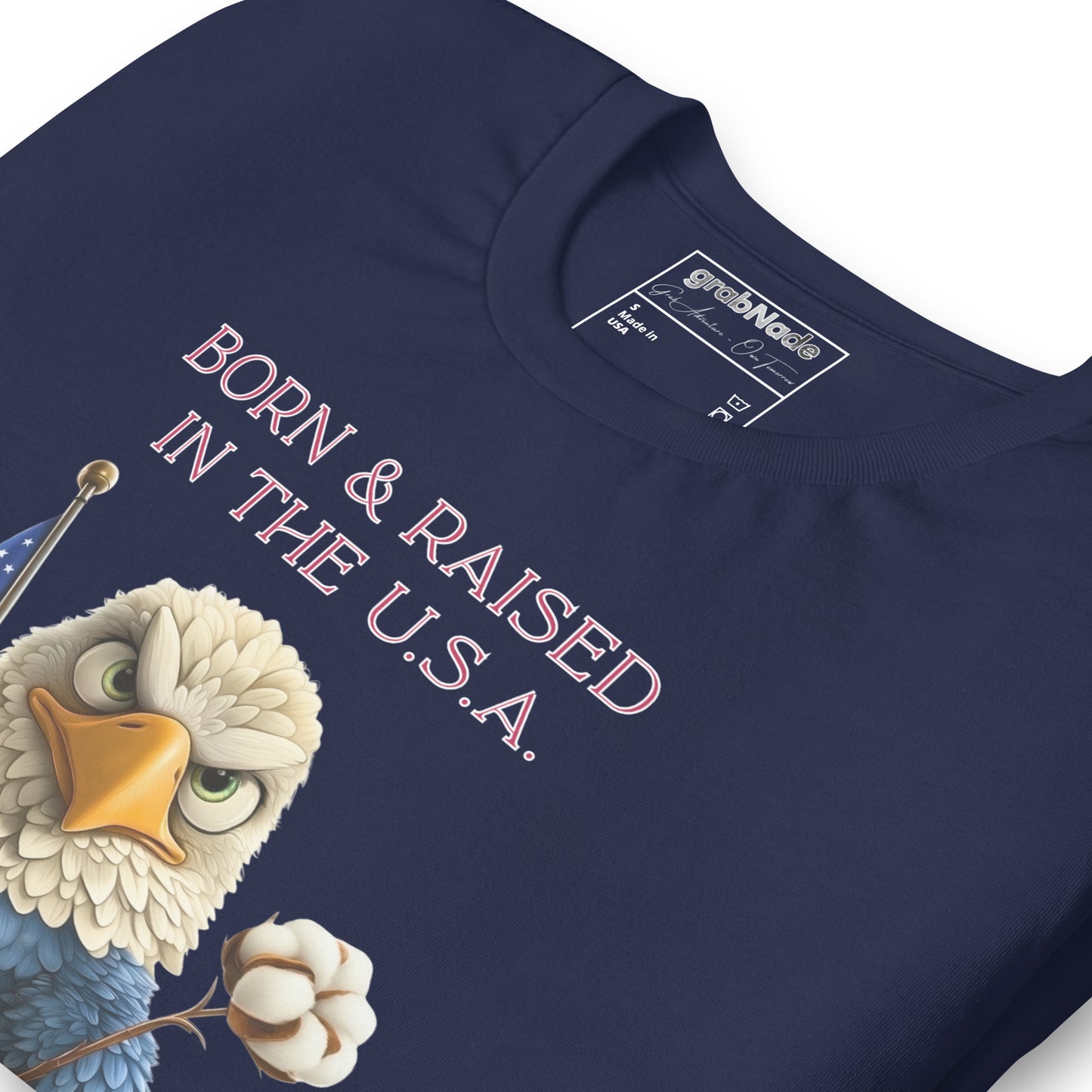 Product mockup. Close-up of navy blue T-shirt with a cartoon bald eagle holding a cotton flower and American flag, text "BORN & RAISED IN THE U.S.A."