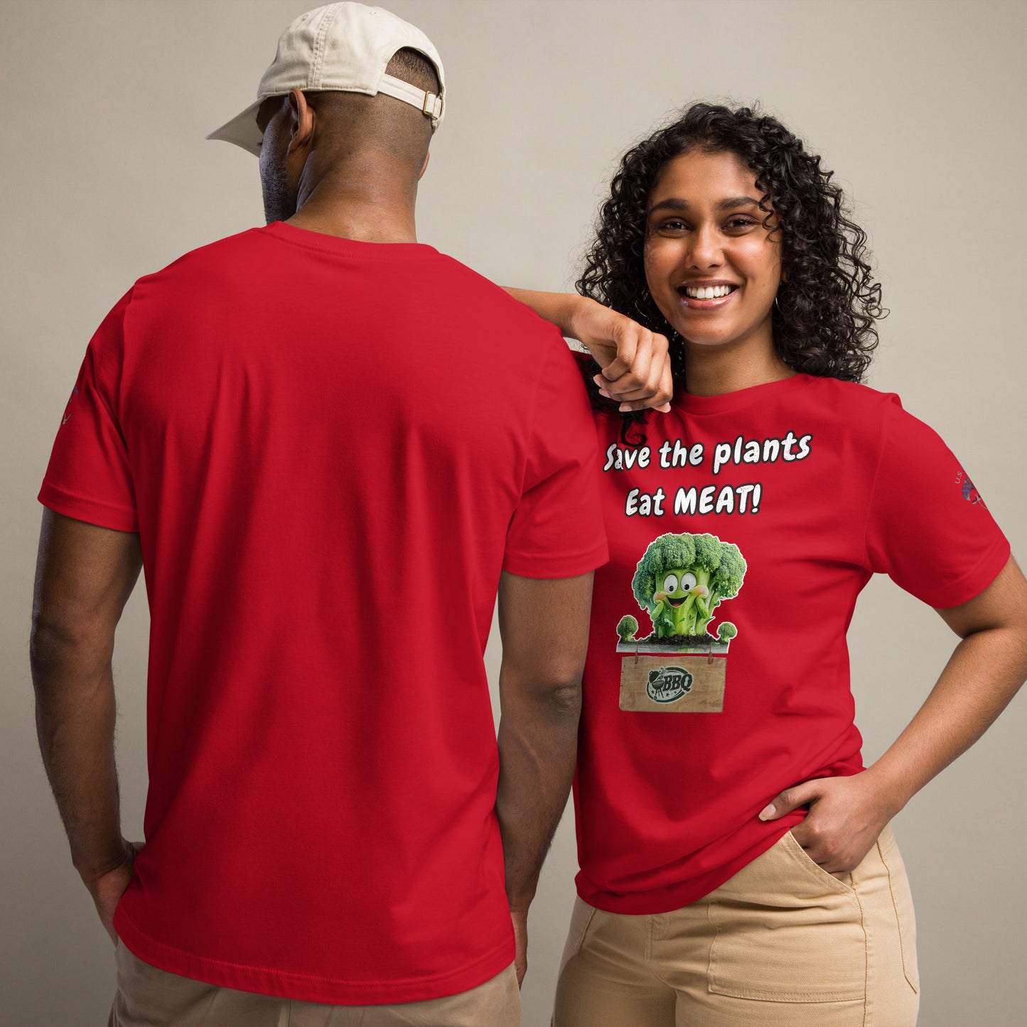 Product mockup. A man and a woman posing together, both in beige pants; the woman is showing the back of a red T-shirt with a cartoon broccoli and “Save the plants Eat MEAT!” text.