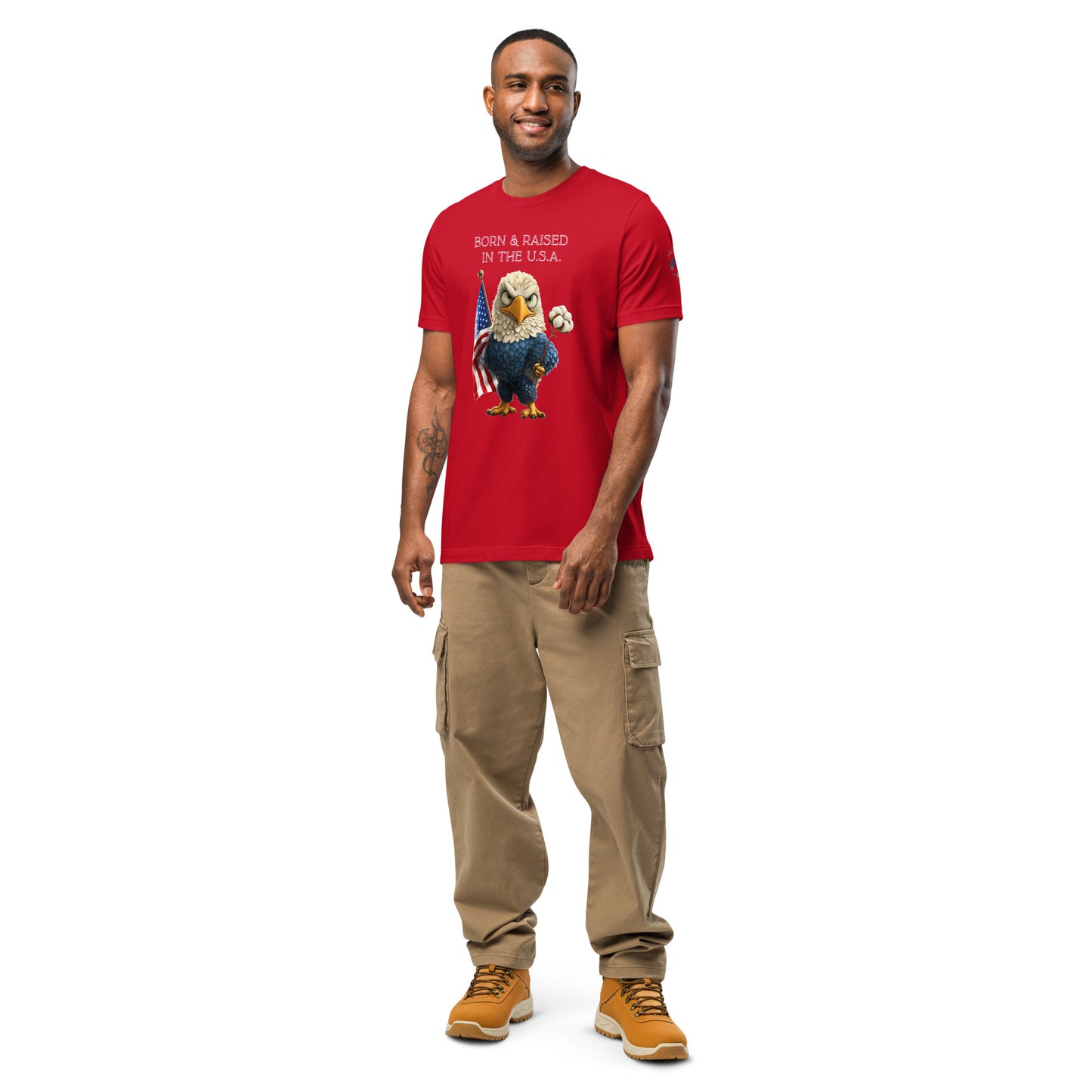 Product mockup. Person standing wearing red T-shirt displaying stylized eagle holding American flag and cotton stem with 'Born & Raised in the U.S.A.' text, khaki cargo pants, and tan boots.