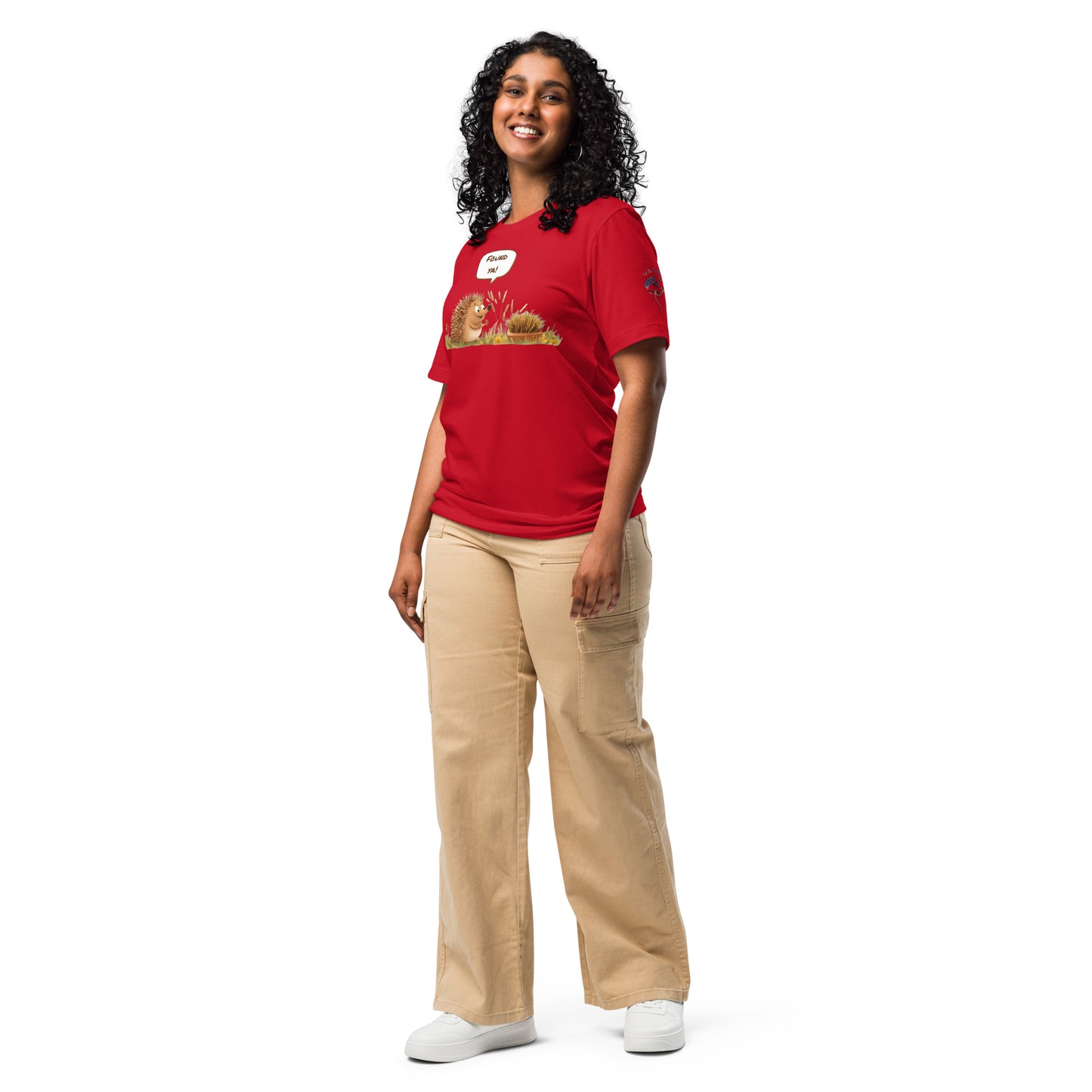 Product mockup. A woman dressed in beige pants and a red T-shirt featuring two cartoon hedgehogs in the grass, one exclaiming "Found ya!" in a full-body pose.