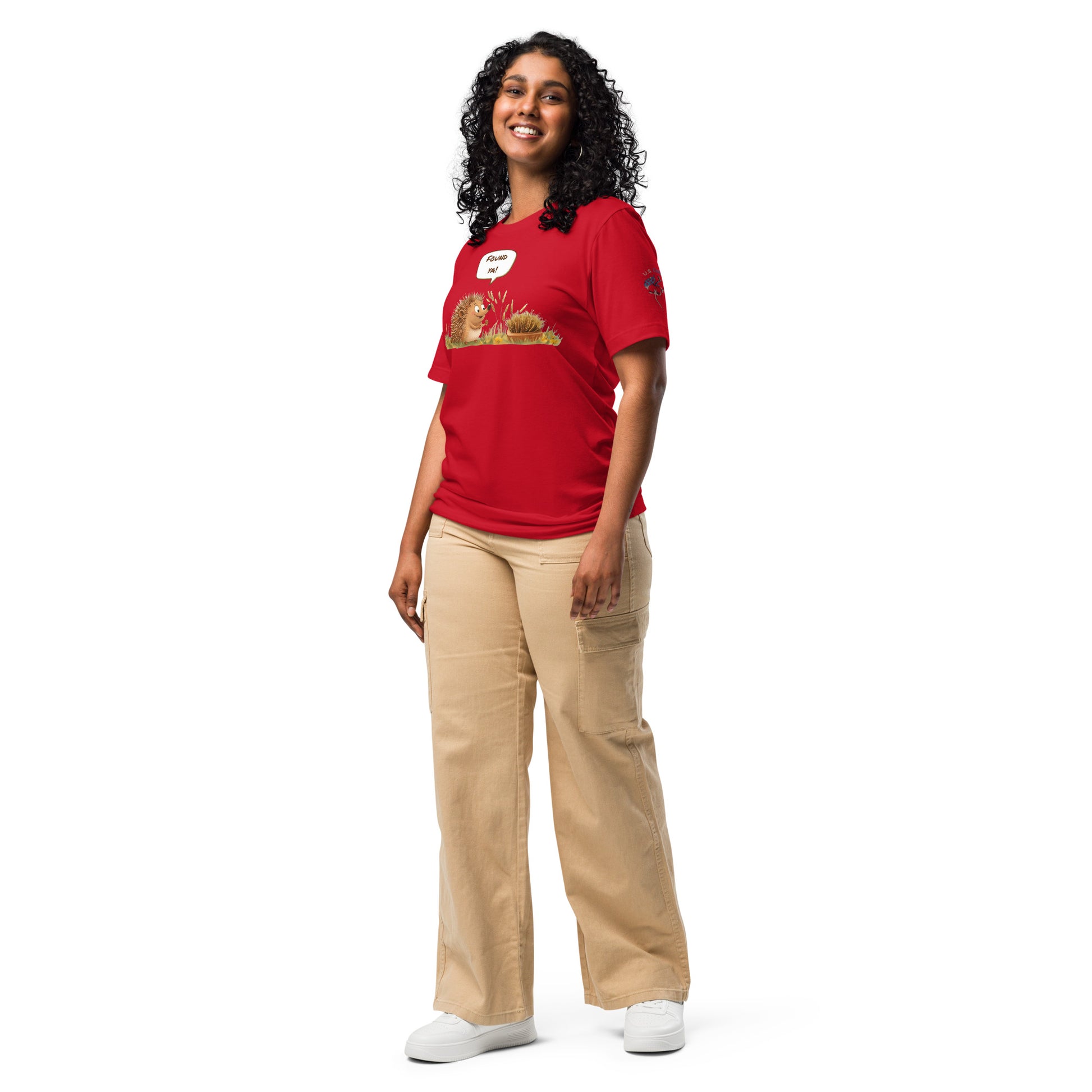 Product mockup. A woman dressed in beige pants and a red T-shirt featuring two cartoon hedgehogs in the grass, one exclaiming "Found ya!" in a full-body pose.