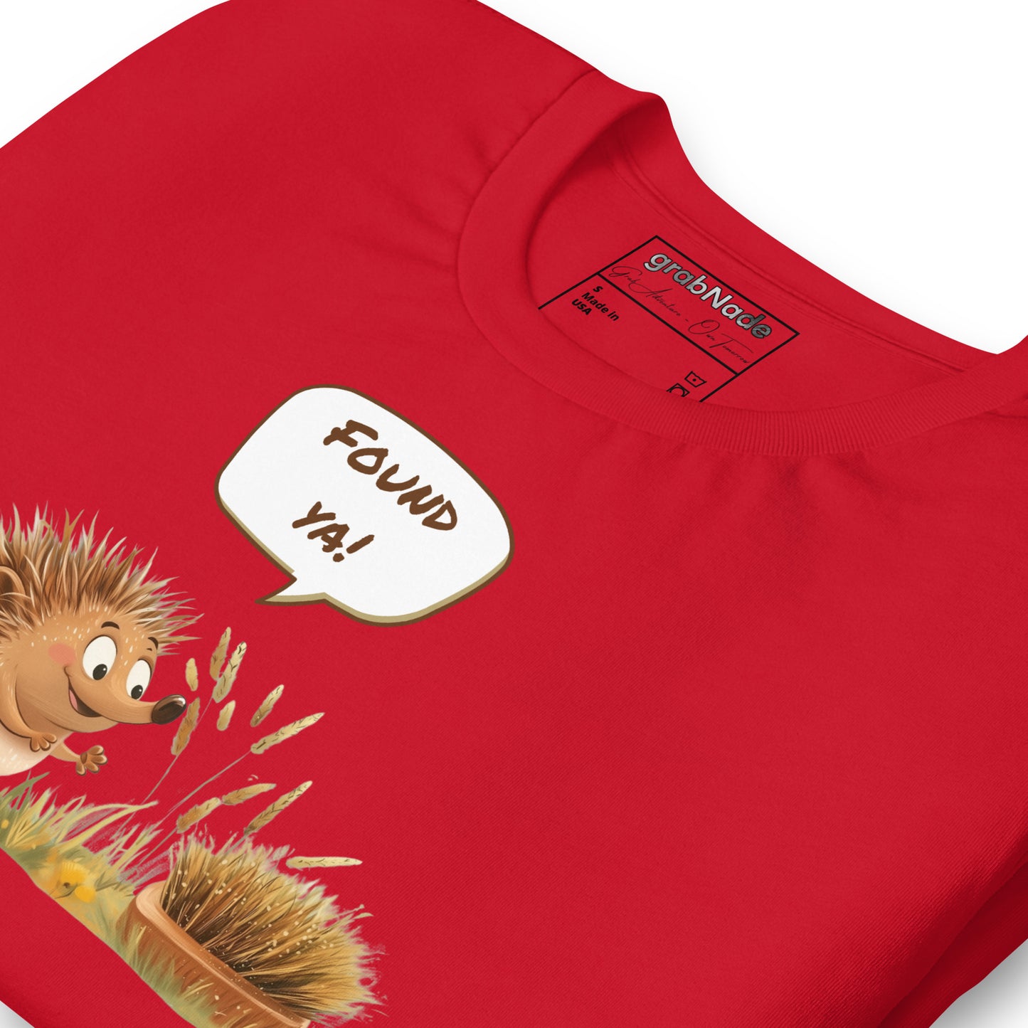 Product mockup. Close-up of a red T-shirt featuring a smiling cartoon hedgehog exclaiming “Found ya!” toward a brush disguised as a hedgehog in grass.