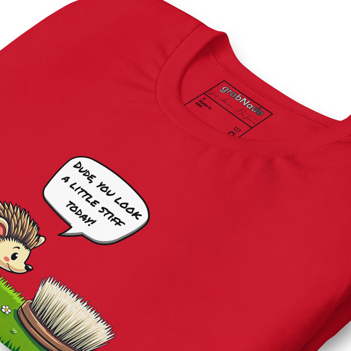 Product mockup. Close-up of a red T-shirt with a cartoon hedgehog saying, “DUDE, YOU LOOK A LITTLE STIFF TODAY!” to a round brush in grass.